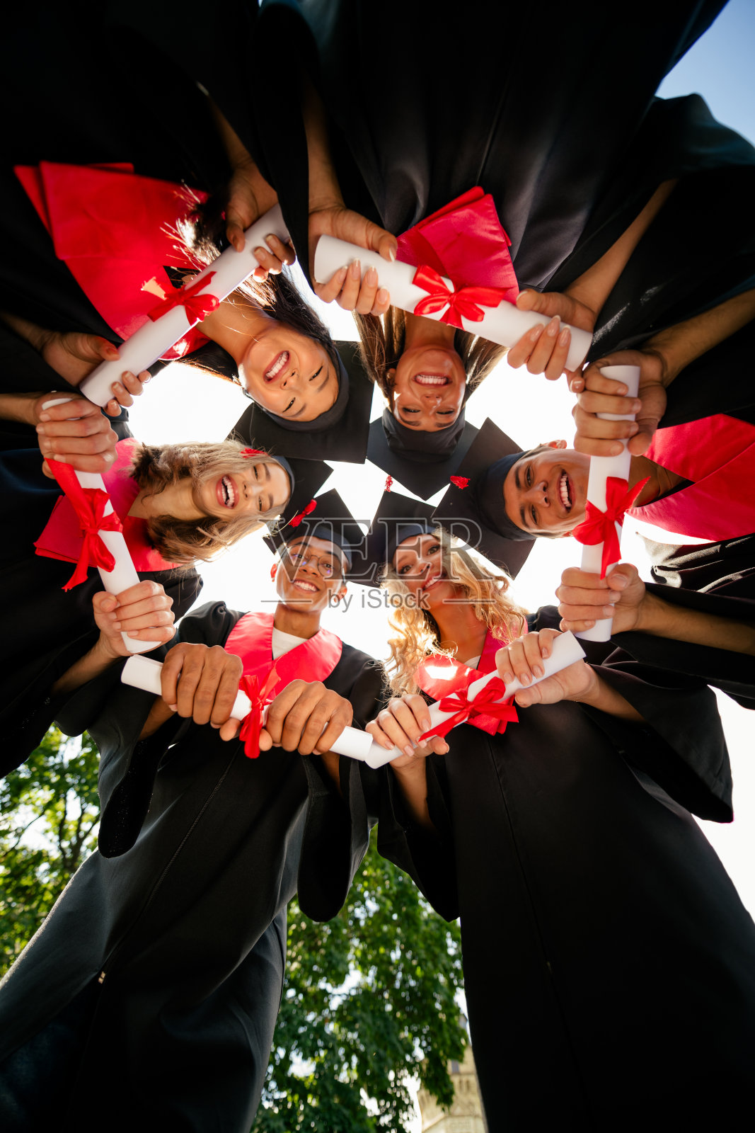 低角度视图的学生群体，露齿而笑，手持文凭，围成一圈，戴着学位帽，穿着毕业袍，户外场景。照片摄影图片