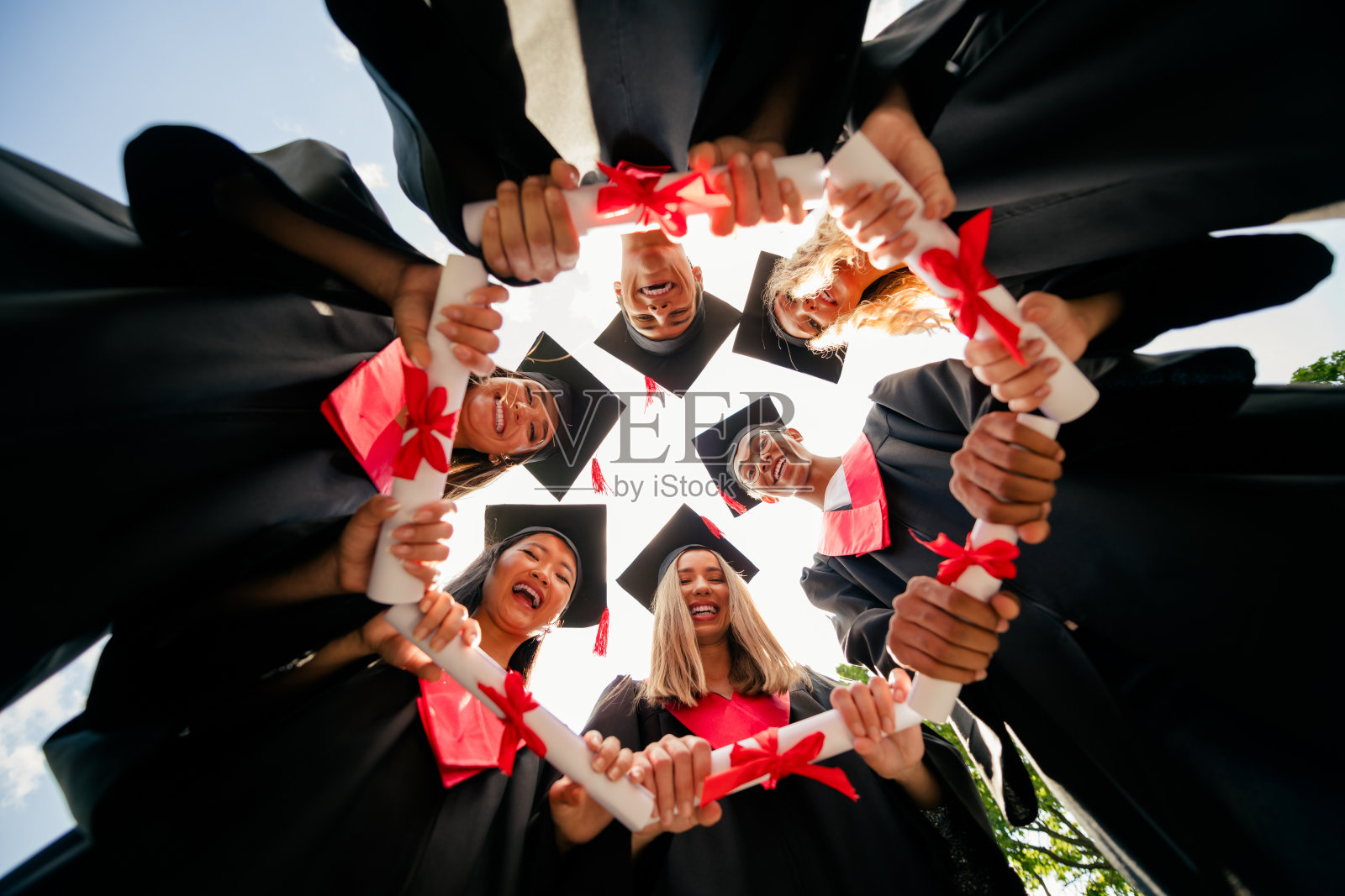 低角度视角的学生群体，手持文凭，围成圆圈，穿着学士服和学位帽，在户外庆祝毕业。照片摄影图片