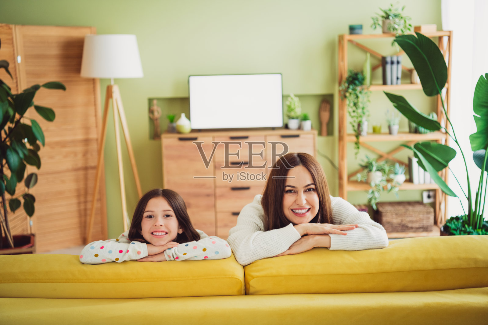 微笑的年轻母亲和女儿在明亮的客厅里放松，坐在黄色沙发上，周围充满自然光。照片摄影图片