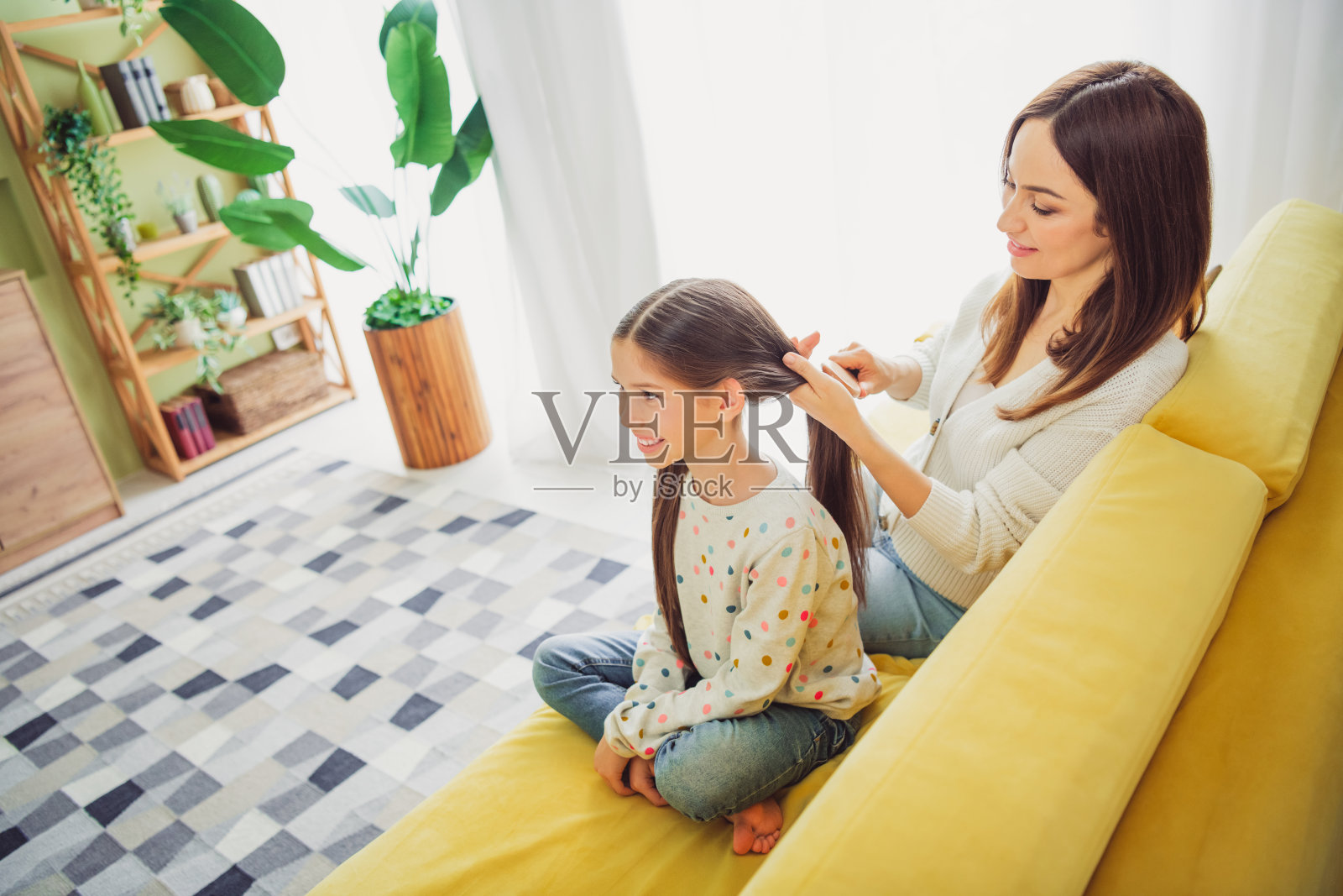 母亲在现代明亮的客厅里坐在黄色沙发上为女儿编辫子，分享着愉快的亲子时光。照片摄影图片