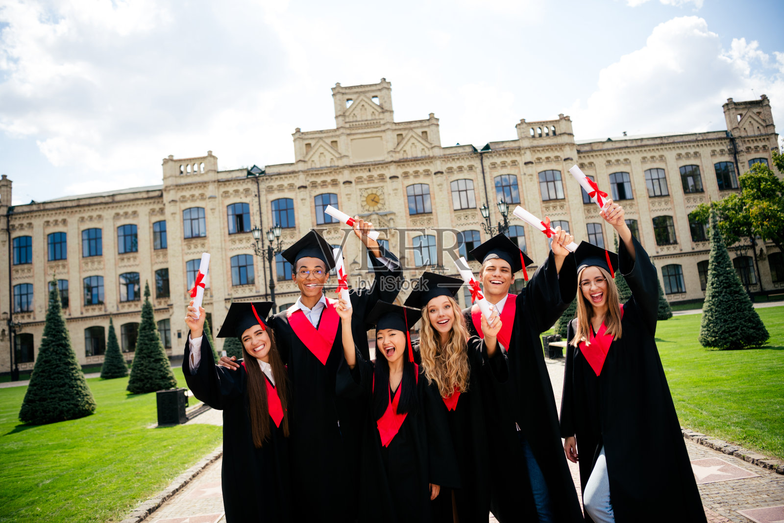 公司同学们在户外举起文凭，戴着学位帽和毕业袍，合影于大学建筑前。照片摄影图片