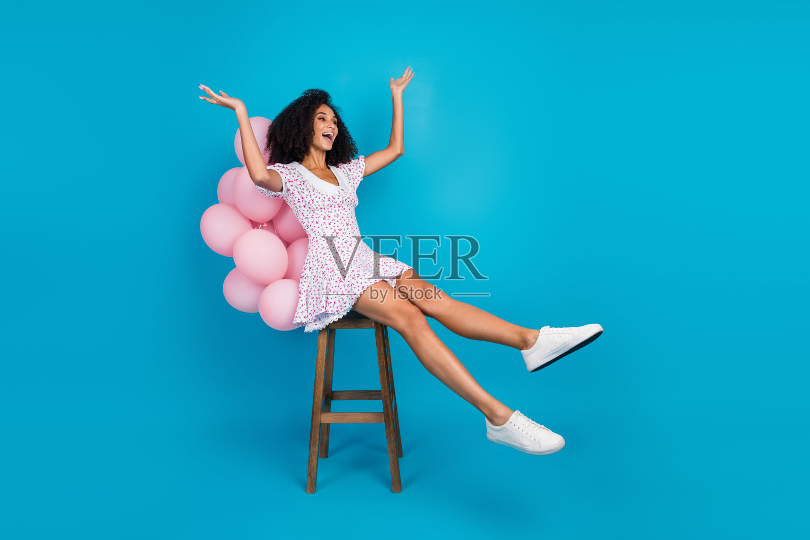 穿着印花裙的时尚年轻女性坐在凳子上，面前有粉色气球，背景是明亮的蓝色。照片摄影图片