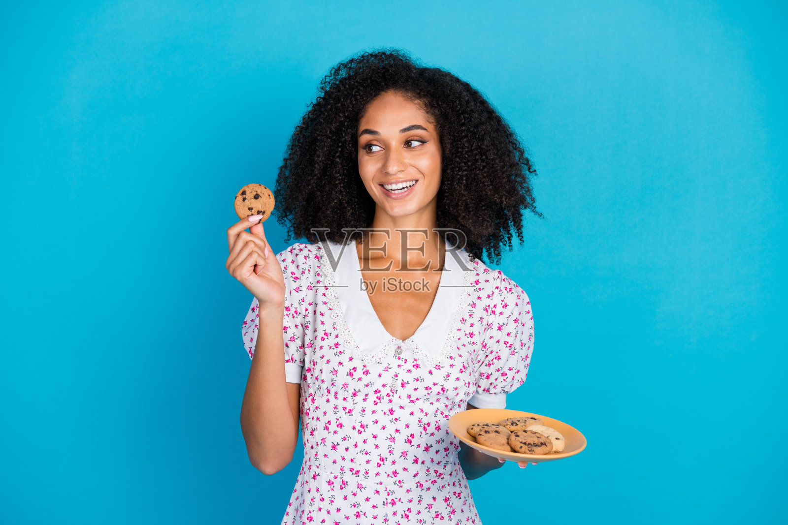 穿着时尚花卉裙的快乐年轻女性在蓝色背景前手持巧克力曲奇饼干照片摄影图片