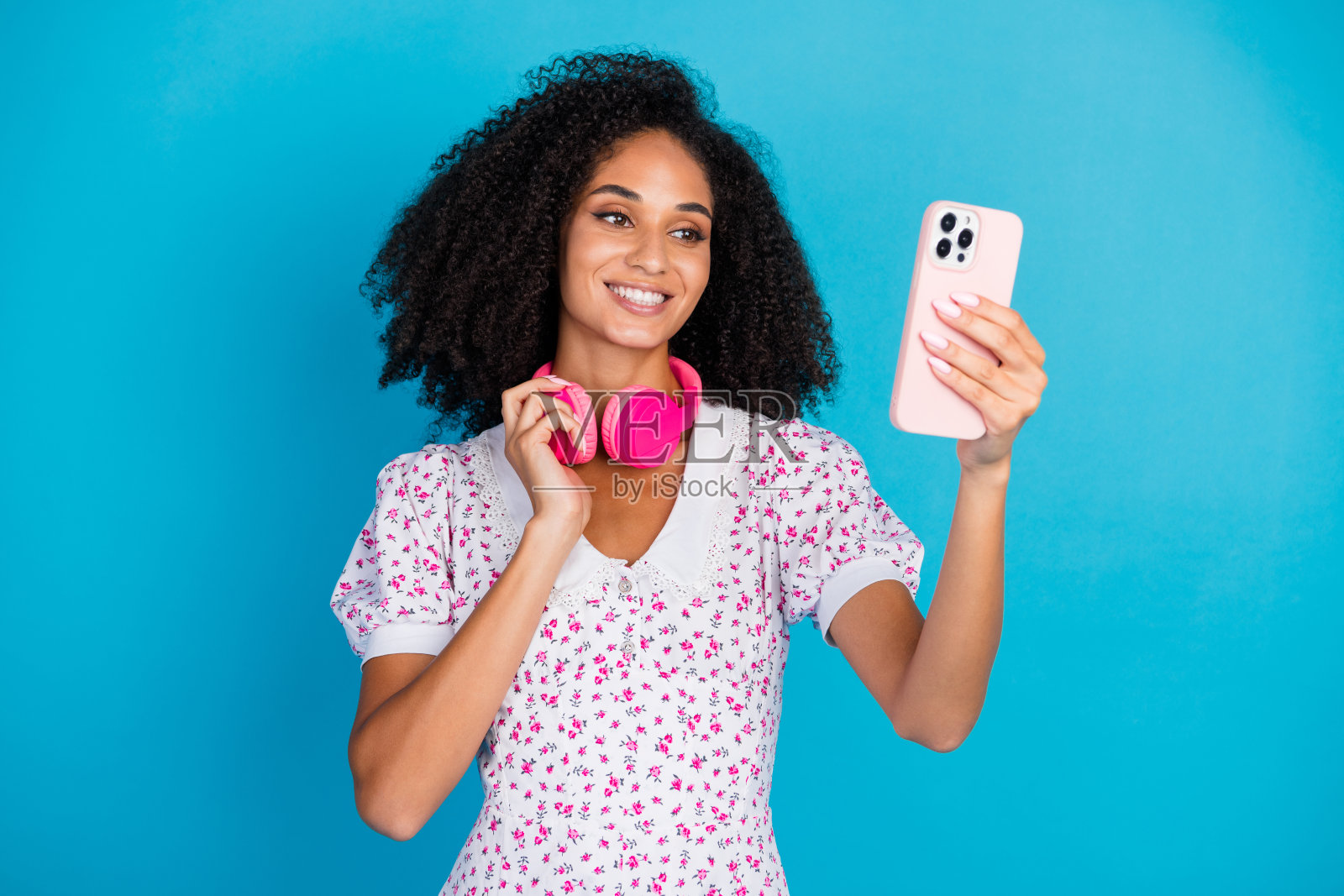 年轻女性卷发，手持智能手机，佩戴粉色耳机，背景为鲜艳的蓝色。照片摄影图片