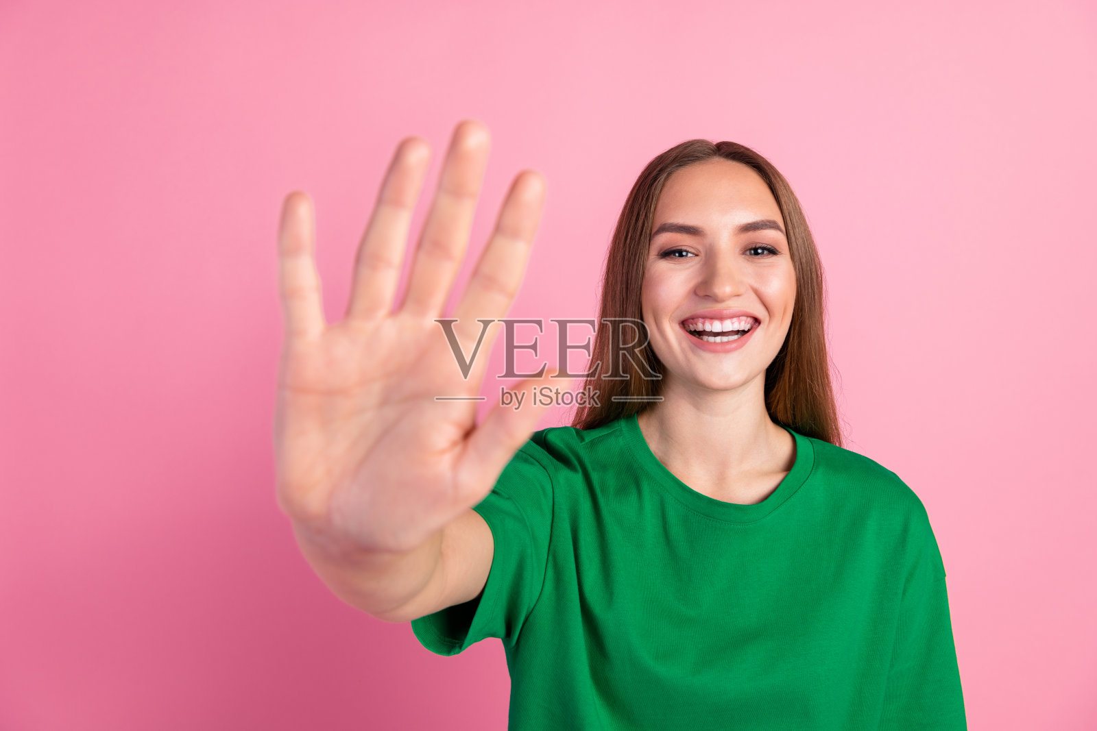 穿着绿色T恤的年轻女性在充满活力的粉色背景前微笑着，愉快地挥手，展现出快乐和温暖。照片摄影图片