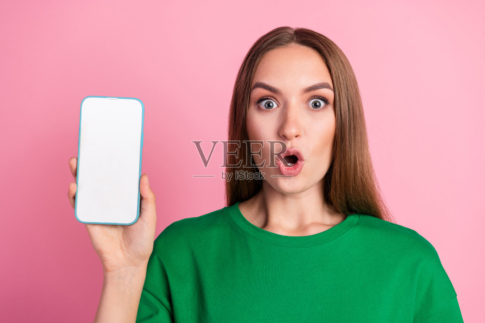 穿着绿色T恤的年轻女性，惊讶地指着智能手机，站在鲜艳的粉色背景前。照片摄影图片