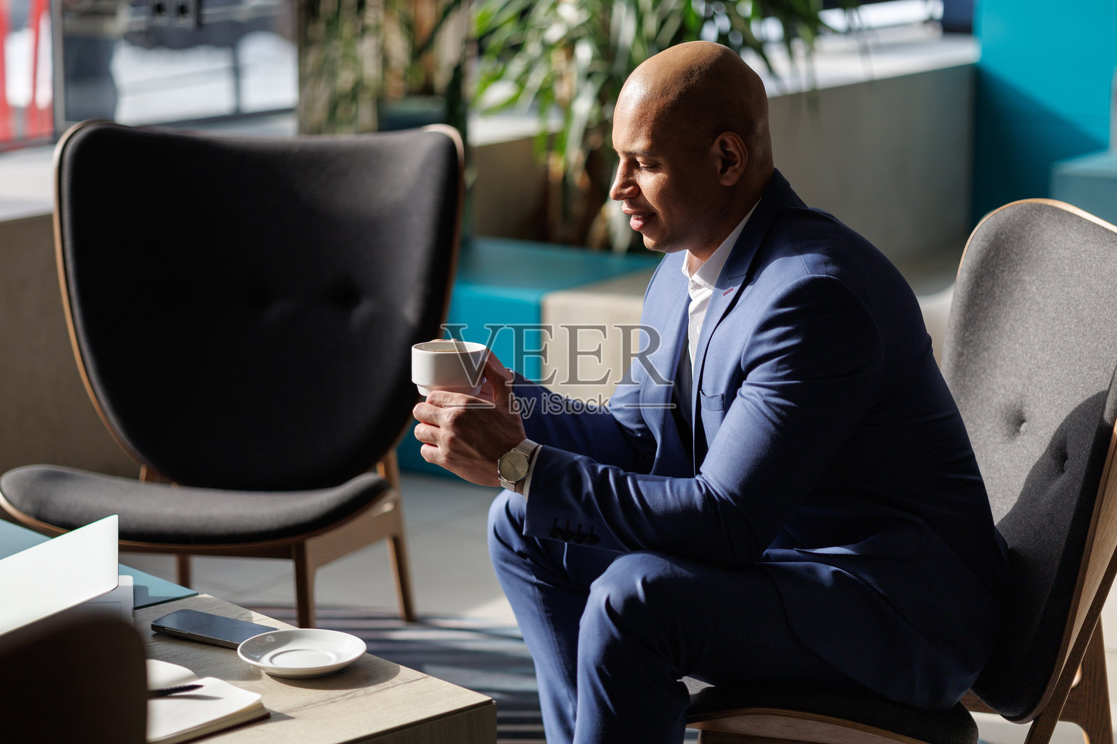 非裔美国商人在现代酒店内手持咖啡，身边有电子设备。照片摄影图片
