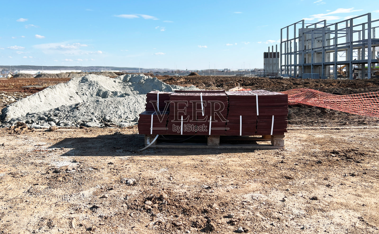 建筑工地展示整齐堆放在托盘上的红色瓷砖，背景中有一部分建成的结构。沙土地面和保护网可见，表明正在进行的施工工作。照片摄影图片