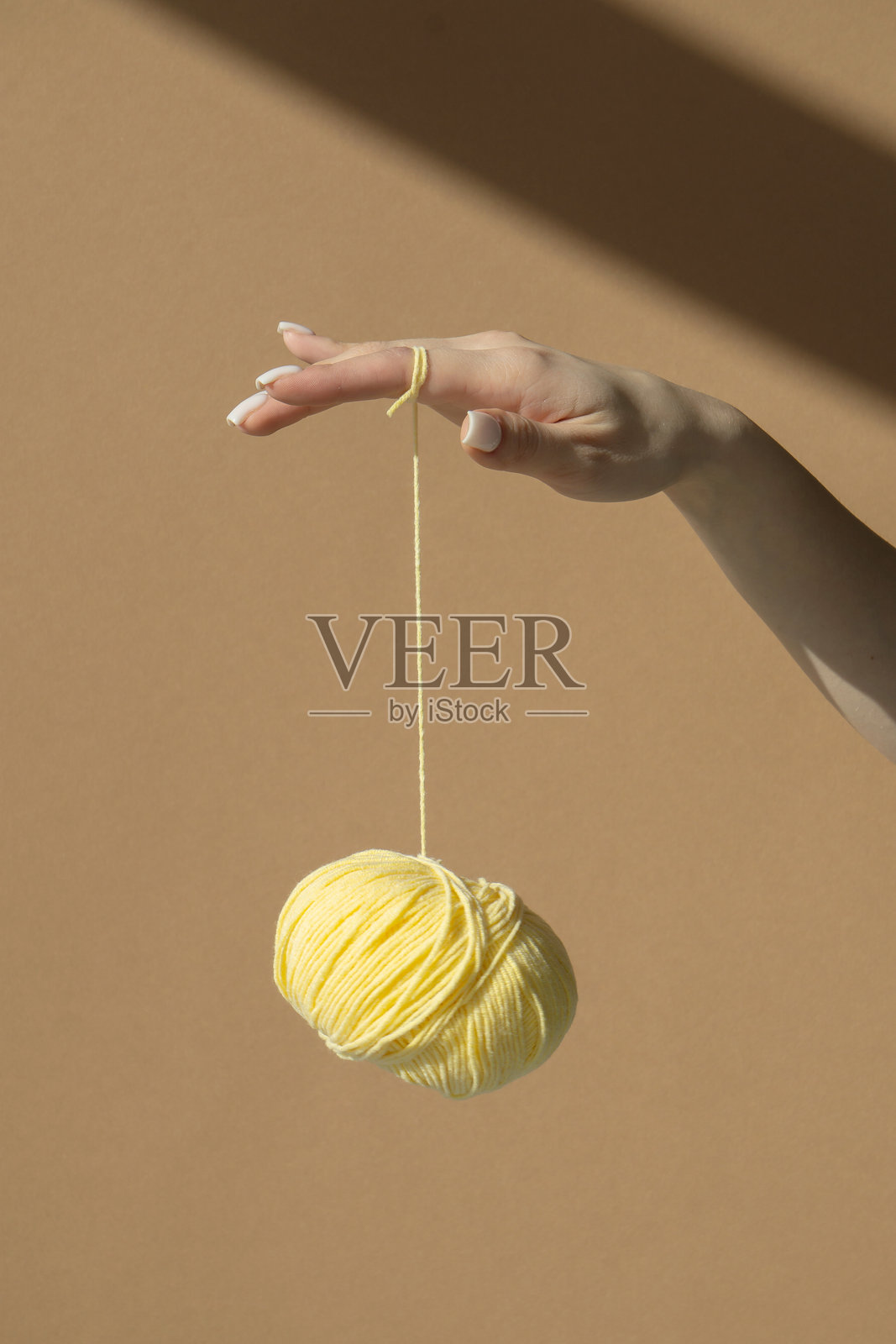 一团黄色的毛线球悬挂在一根优雅女性的白色美甲手指上，背景为棕色，阳光透射而下。创意的样板，竖版框架，留白空间。照片摄影图片
