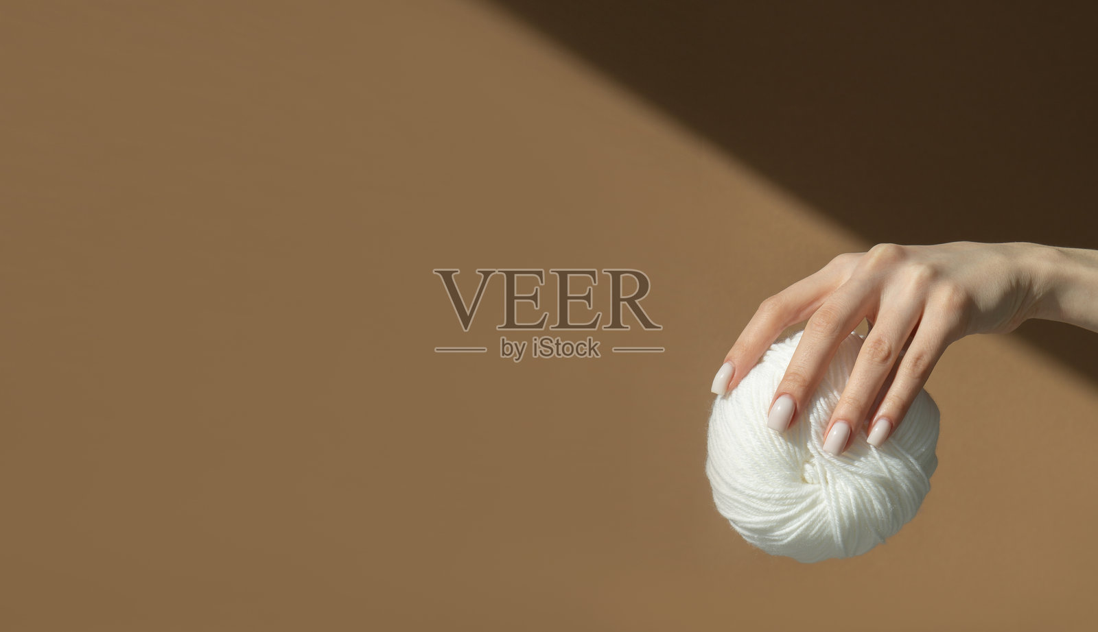 女性手握着一团大的白色毛线球，背景为浅棕色。女人正在握着编织用的毛线。设计师的模板。留有文字空间。照片摄影图片