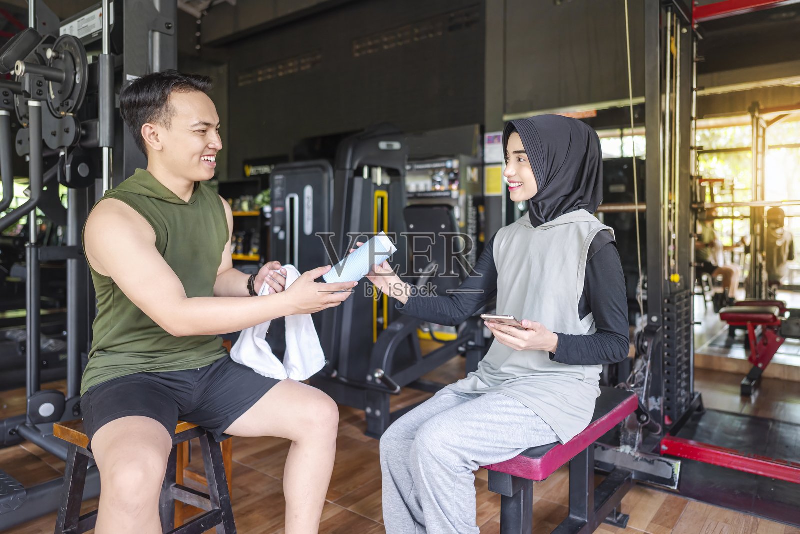 东南亚印尼男性私人教练在健身房里将运动塑料水瓶递给阿拉伯中东穆斯林女性。健身锻炼与健康生活方式的概念照片摄影图片