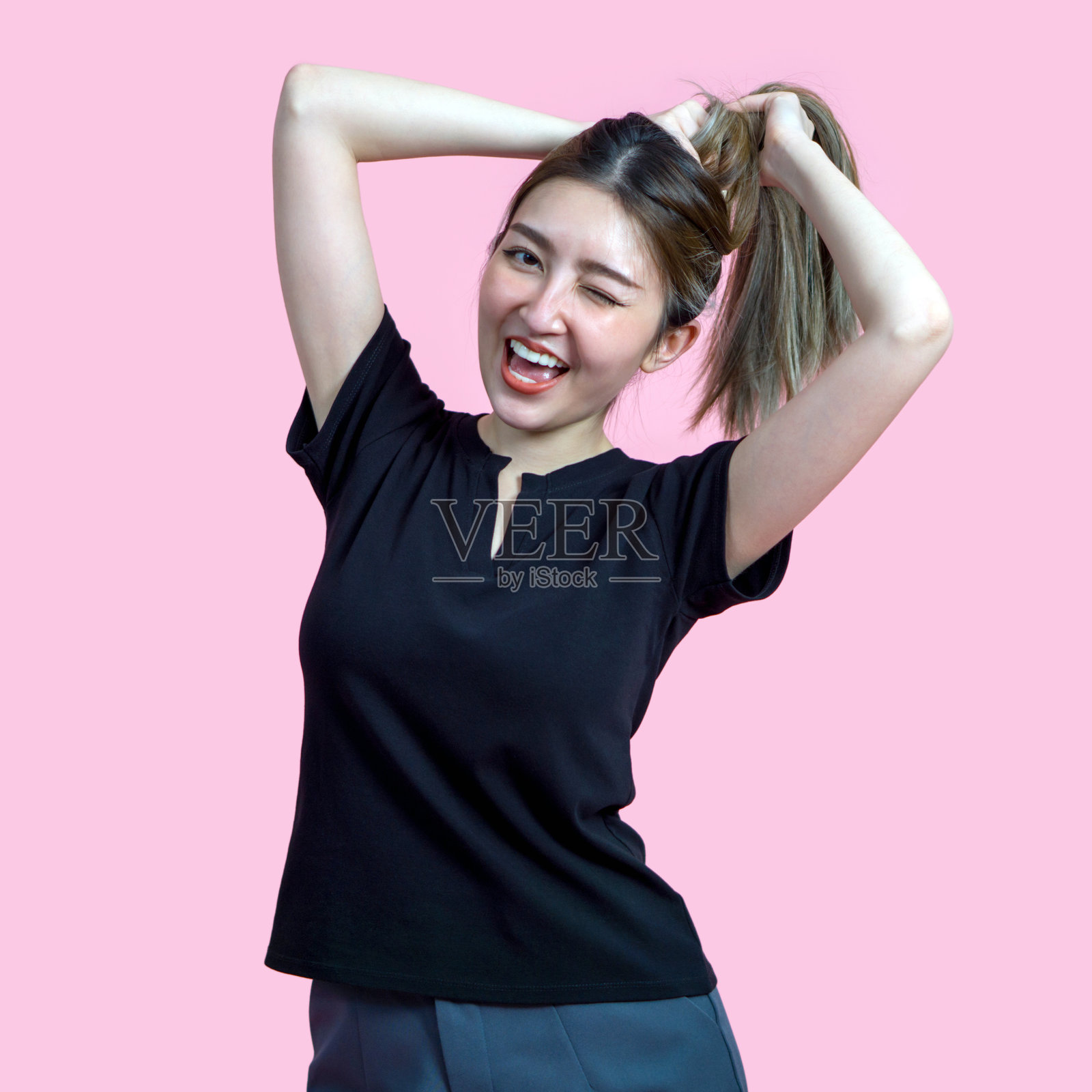 一位年轻女性微笑并眨眼，同时将头发扎成马尾。她穿着黑色T恤，背景是柔和的粉色，营造出活泼的氛围。照片摄影图片