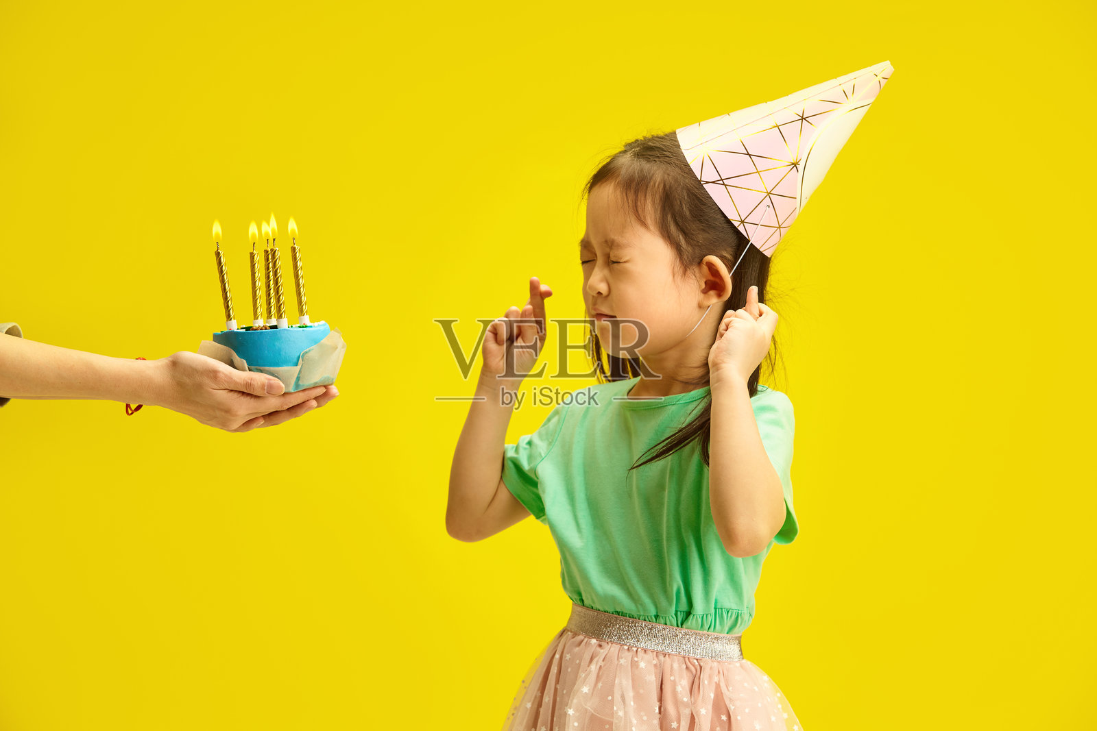 小女孩在黄色背景前许愿，闭着眼睛交叉手指，戴着派对帽，表达对愿望实现的希望。照片摄影图片