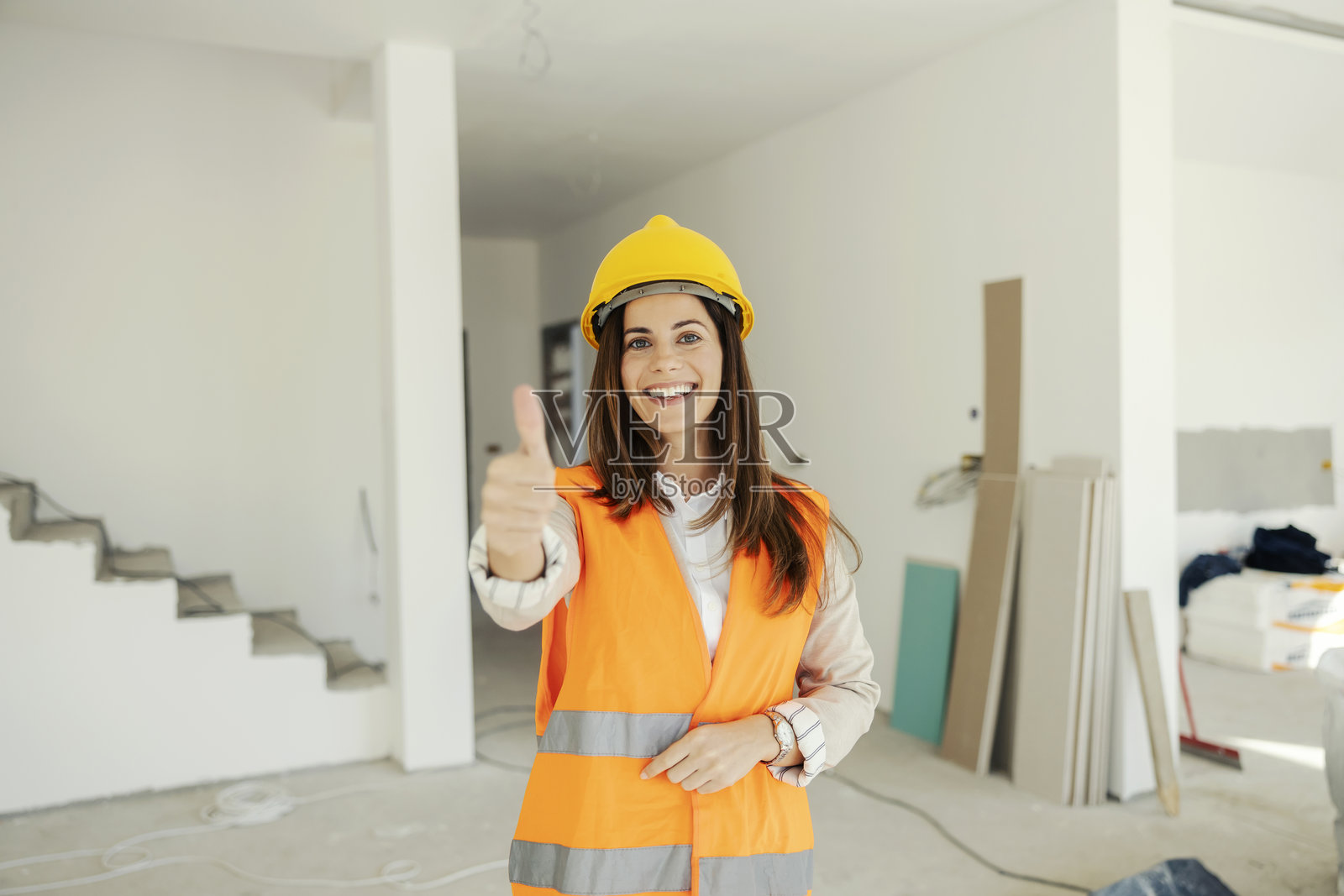 成功的年轻女性建筑师穿着防护服在工地上对着镜头竖起大拇指。照片摄影图片