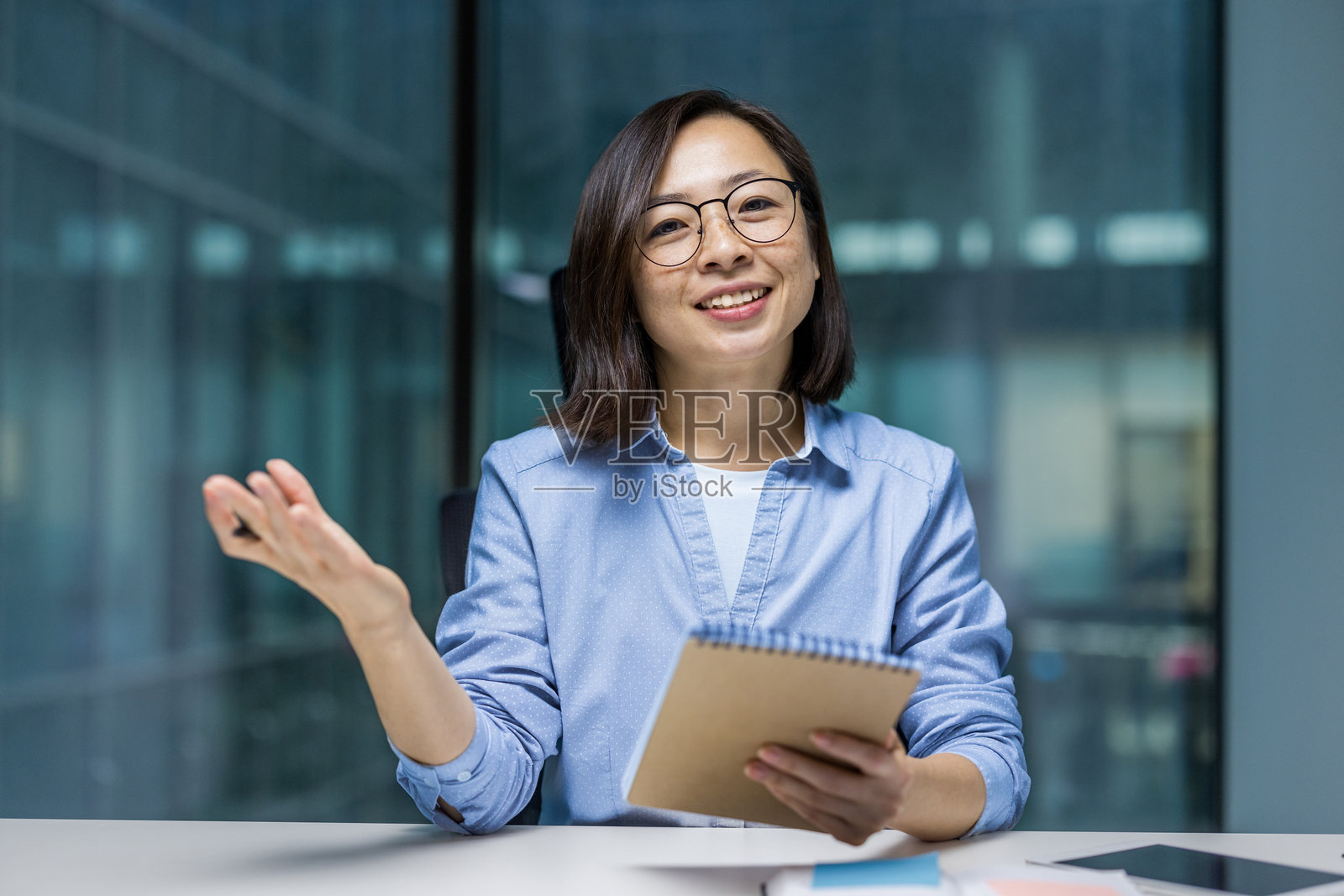 网络摄像头视角，一位女性手持笔记本，微笑着看向镜头。亚洲女性在在线商务会议中，记录数据并在办公室内学习。照片摄影图片