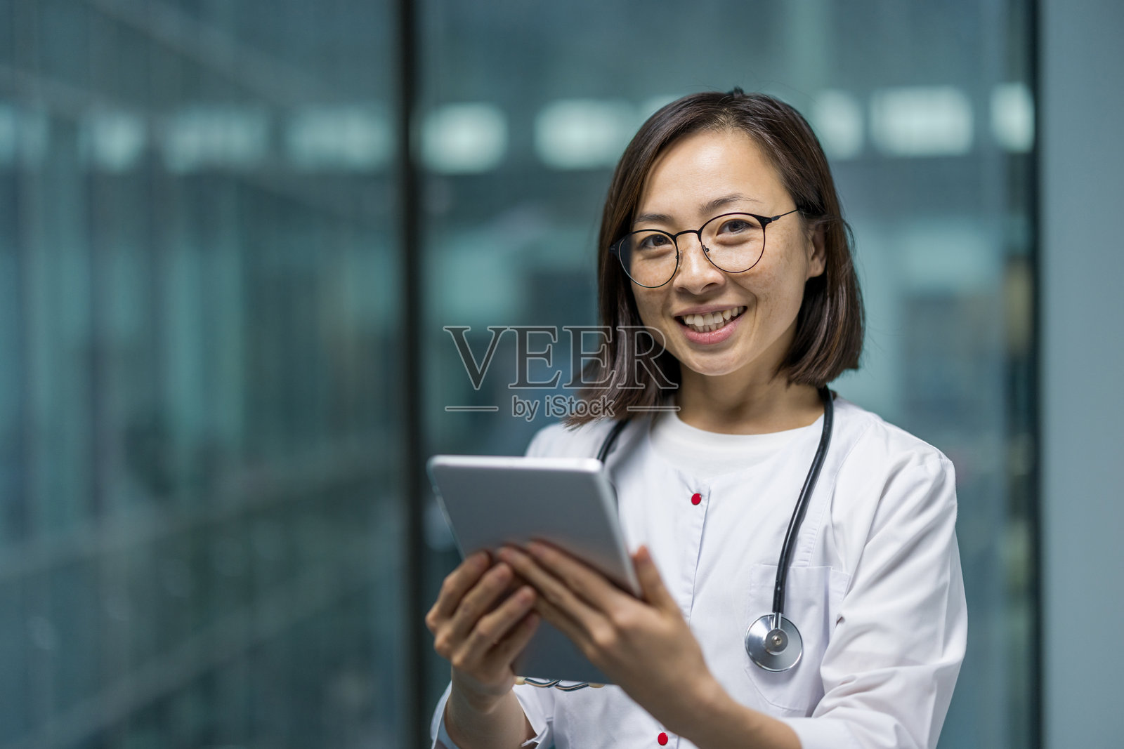 年轻亚洲女医生在办公室内的肖像。女医生穿着白色医师袍，手持平板电脑，微笑着看向镜头，站在窗边。照片摄影图片