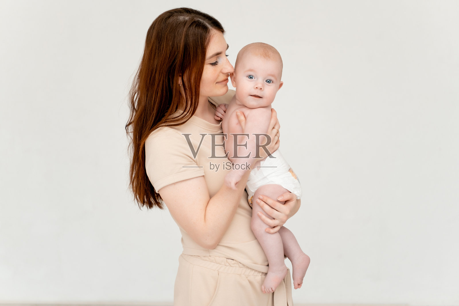 一位年轻母亲在白色背景下轻柔地拥抱她的小孩，旁边留有文字空间，孩子是一个穿着尿布的小女孩，展现了母爱的关怀。照片摄影图片