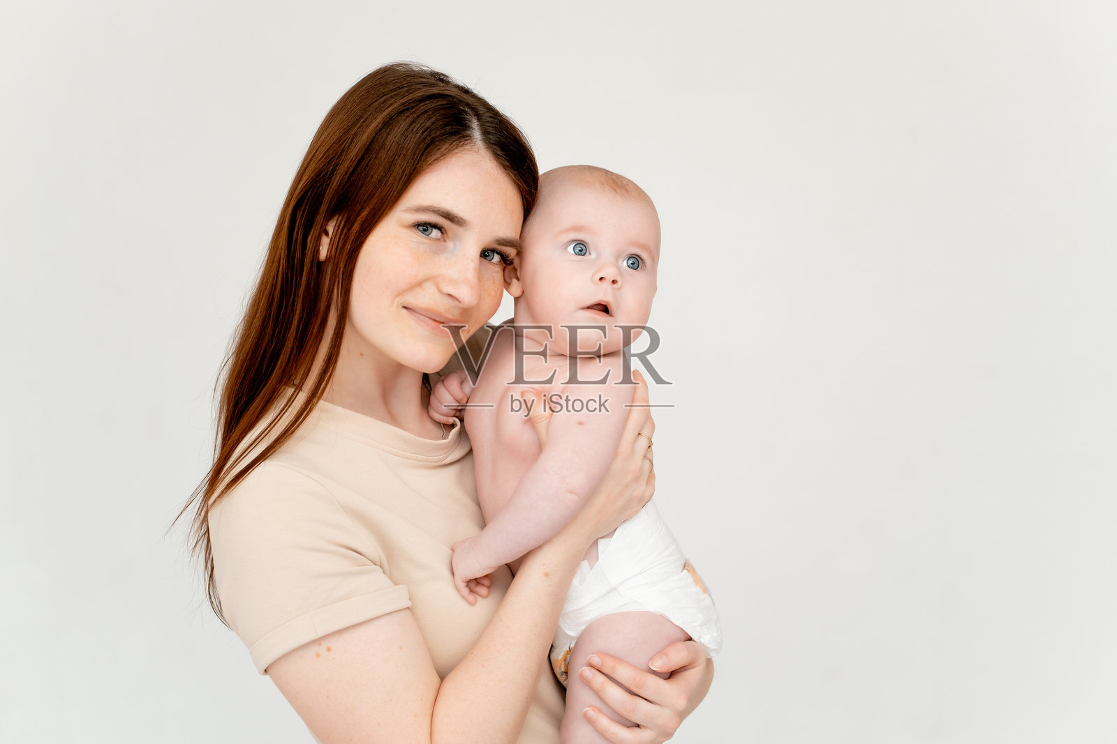 一位年轻母亲在白色背景下温柔地拥抱她的小孩，旁边留有文字空间，孩子是一个穿着尿布的小女孩，展现了母爱的关怀。照片摄影图片