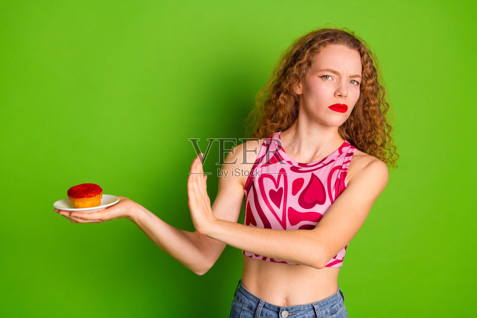 年轻女性一边摇头一边拿着杯子蛋糕，穿着心形图案的上衣，涂着红色口红，背景为绿色。照片摄影图片