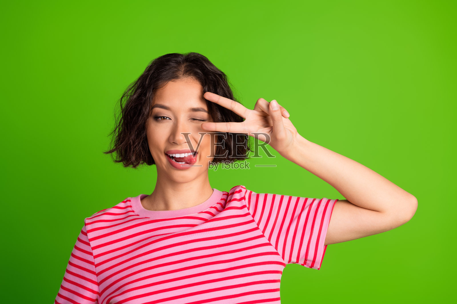 微笑的年轻女性穿着休闲条纹衬衫，做出顽皮的手势，背景是充满活力的绿色，强调乐趣。照片摄影图片