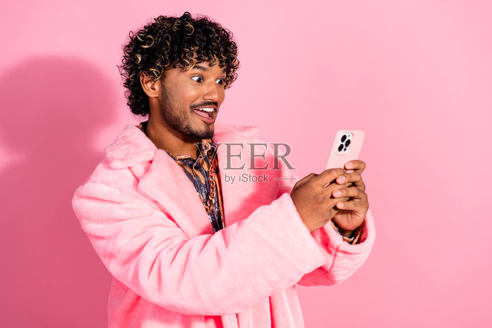 年轻时尚的男士穿着粉色外套，微笑着手持智能手机，背景为明亮的粉色。照片摄影图片