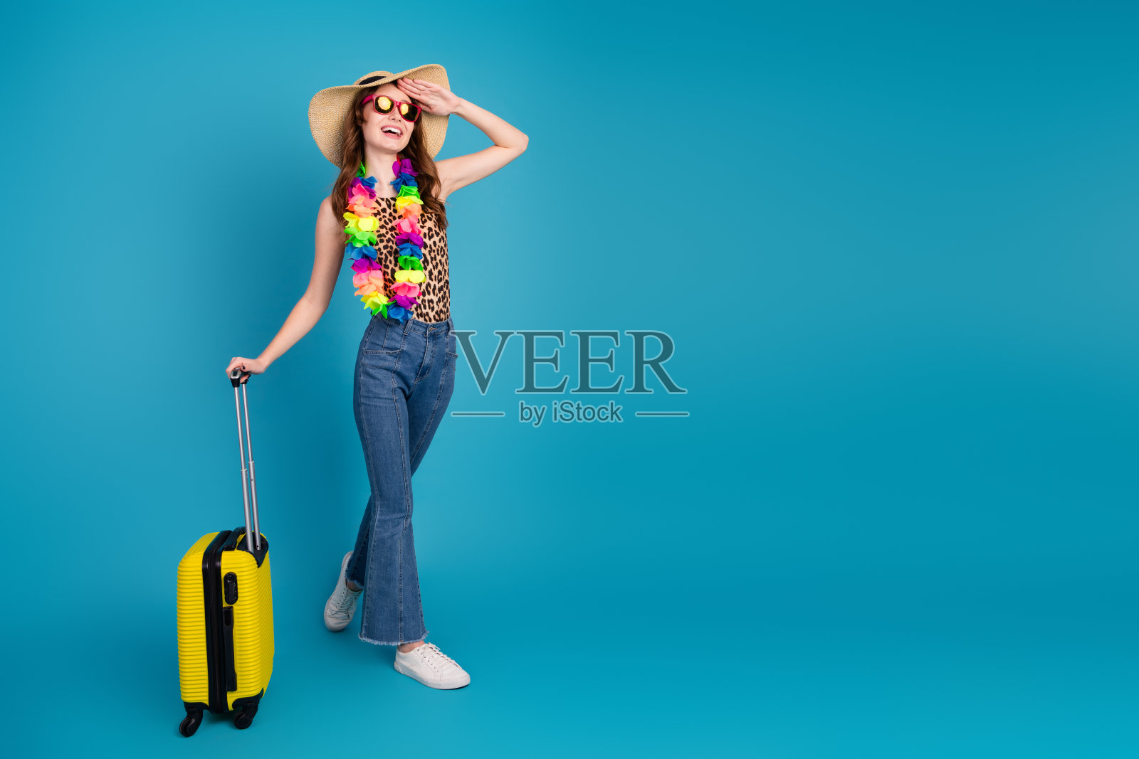 年轻女性穿着夏季服装，手持行李箱在鲜艳的蓝色背景前微笑摆姿势照片摄影图片