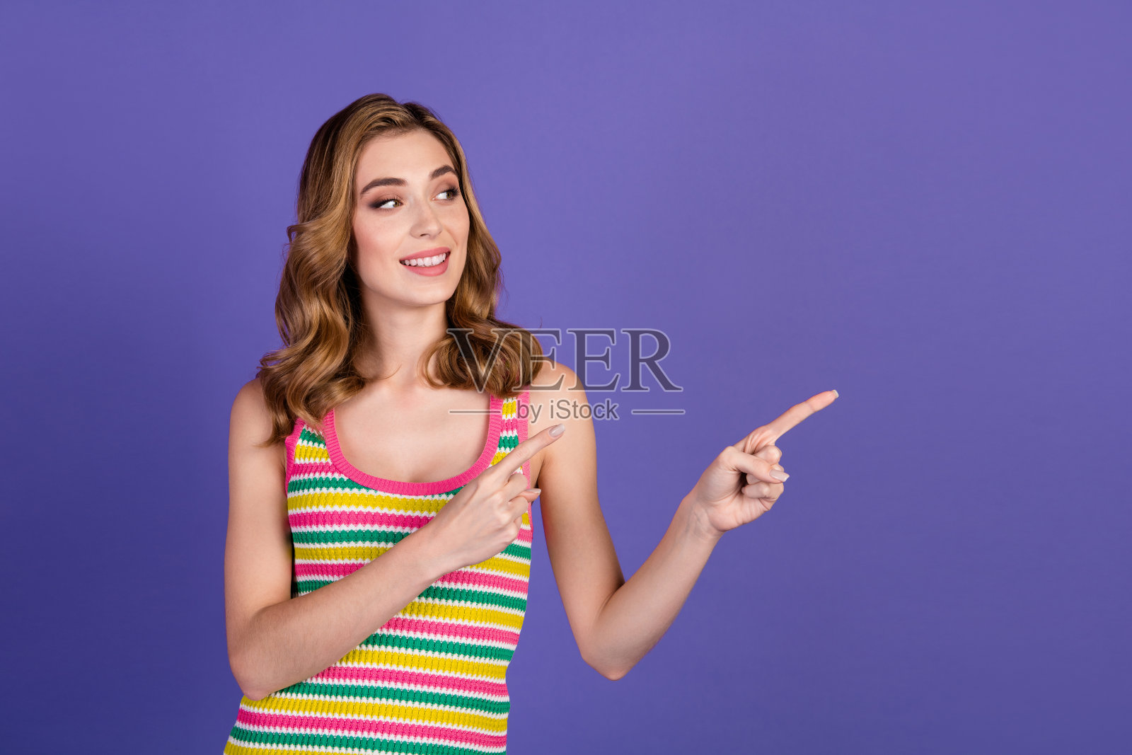 迷人的年轻女性自信地指着，穿着时尚多彩的服装，背景为紫色。照片摄影图片