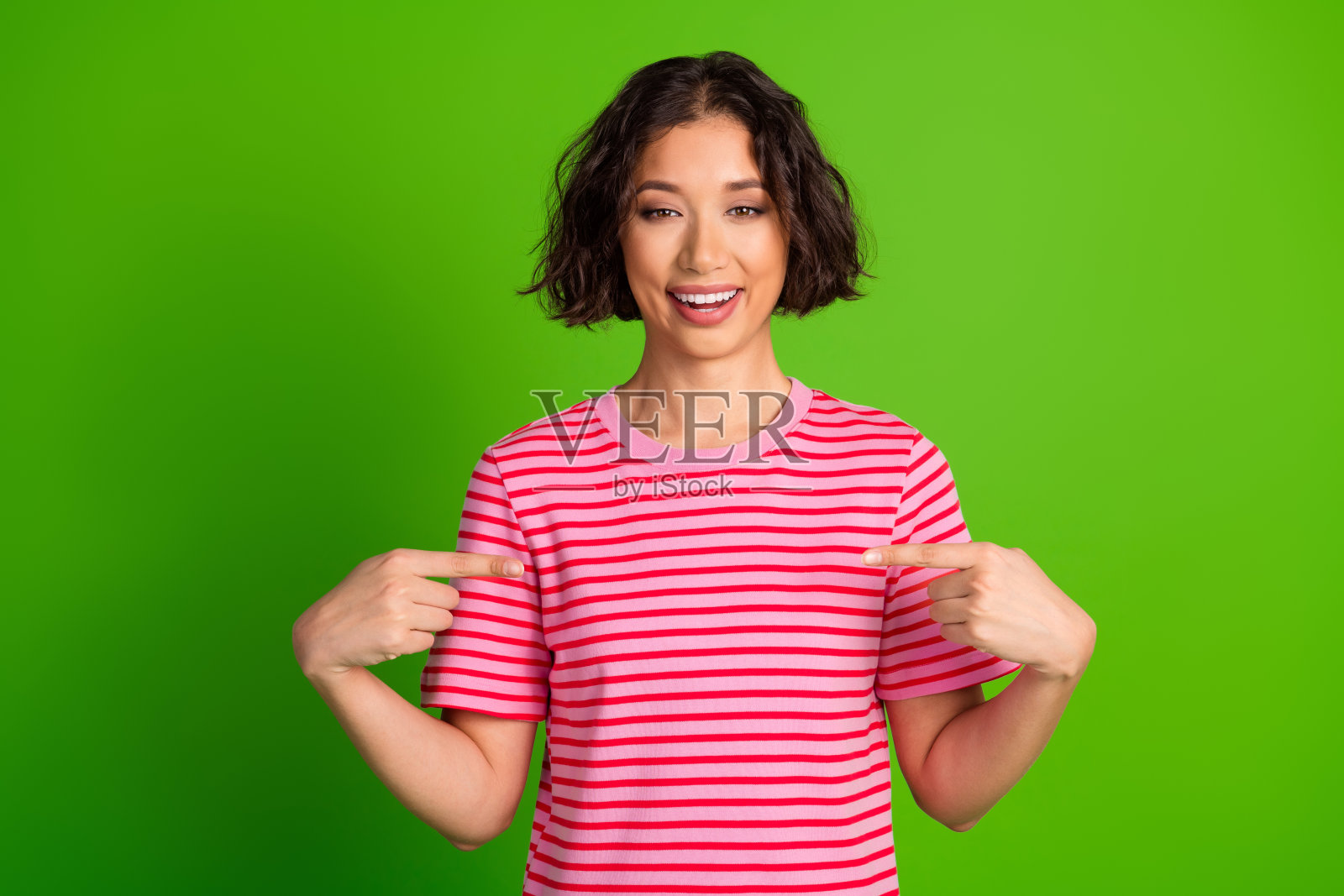 微笑的年轻女性穿着粉色条纹T恤，在充满活力的绿色背景前做手势，适合广告和生活方式展示照片摄影图片