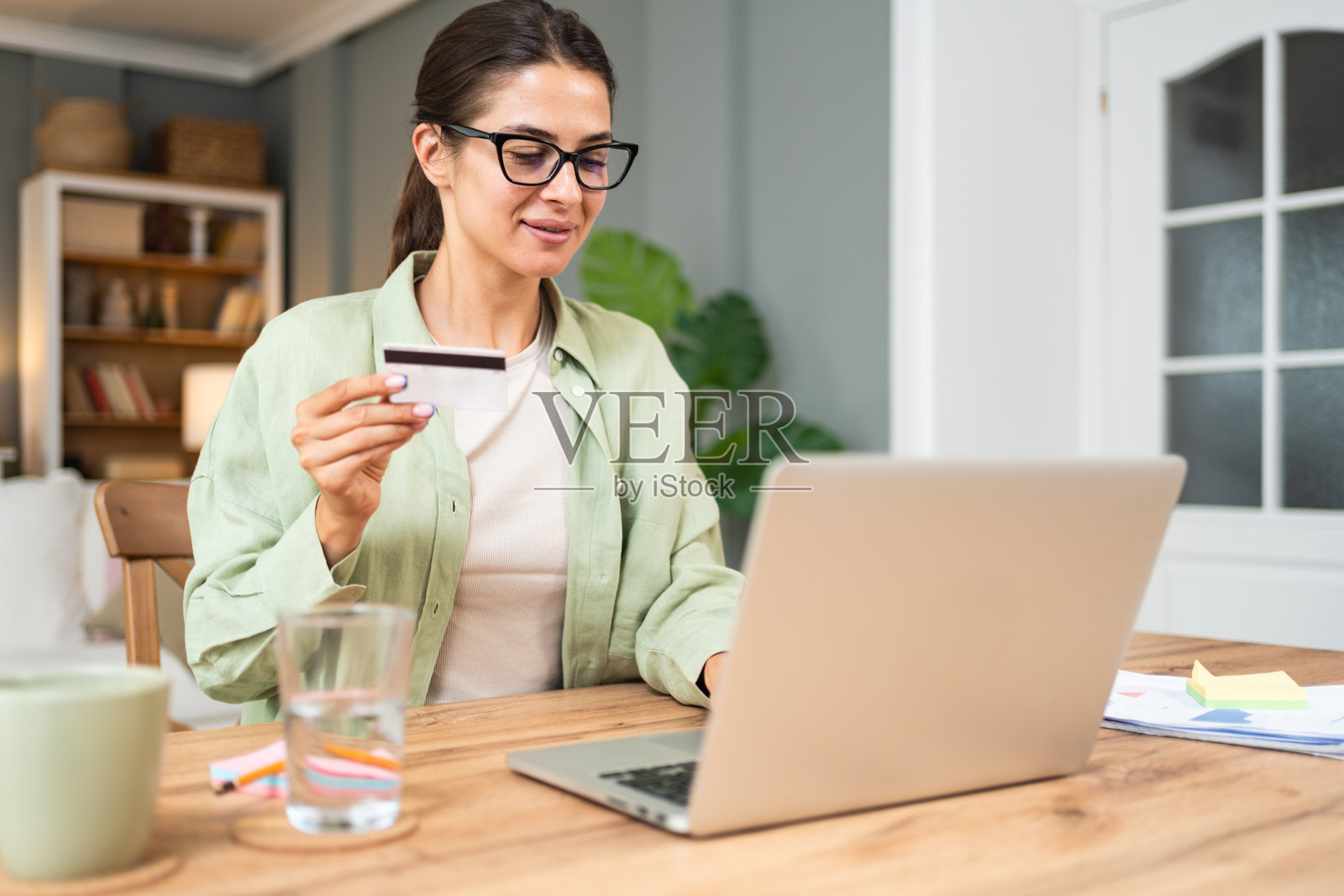 年轻的自由职业女性，坐在公寓的家庭办公室里工作，正在休息，使用银行卡进行网上购物。她很开心，在网上花钱。照片摄影图片