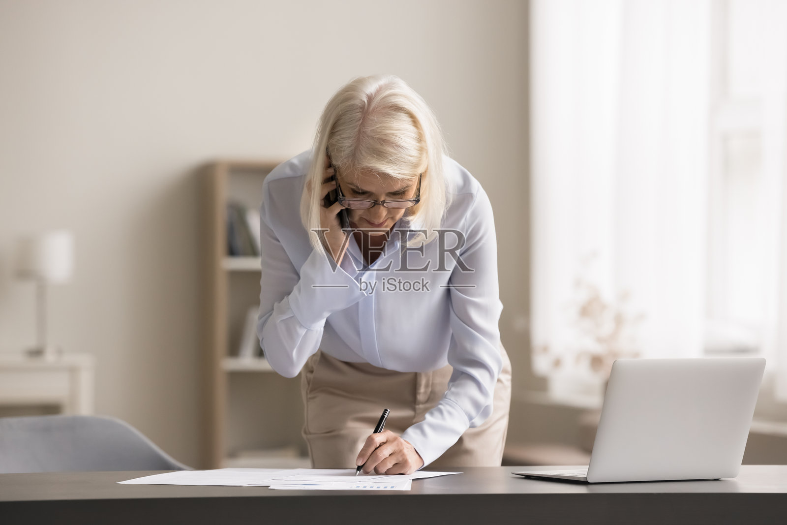 一位年长女性在办公室里使用智能手机通话，同时填写文件。照片摄影图片