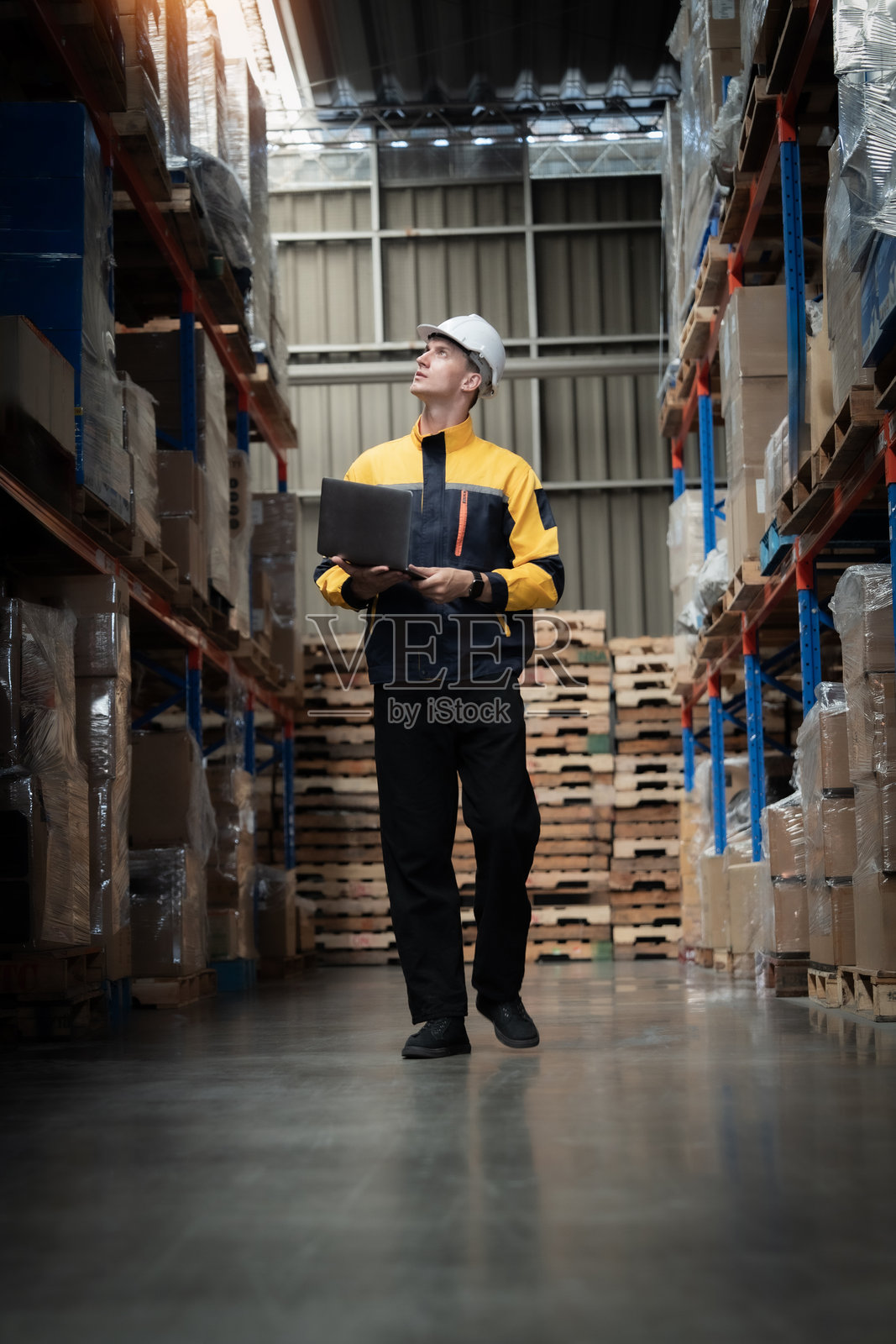 一名穿着黄色和黑色夹克的男子正手持笔记本电脑走过仓库。照片摄影图片