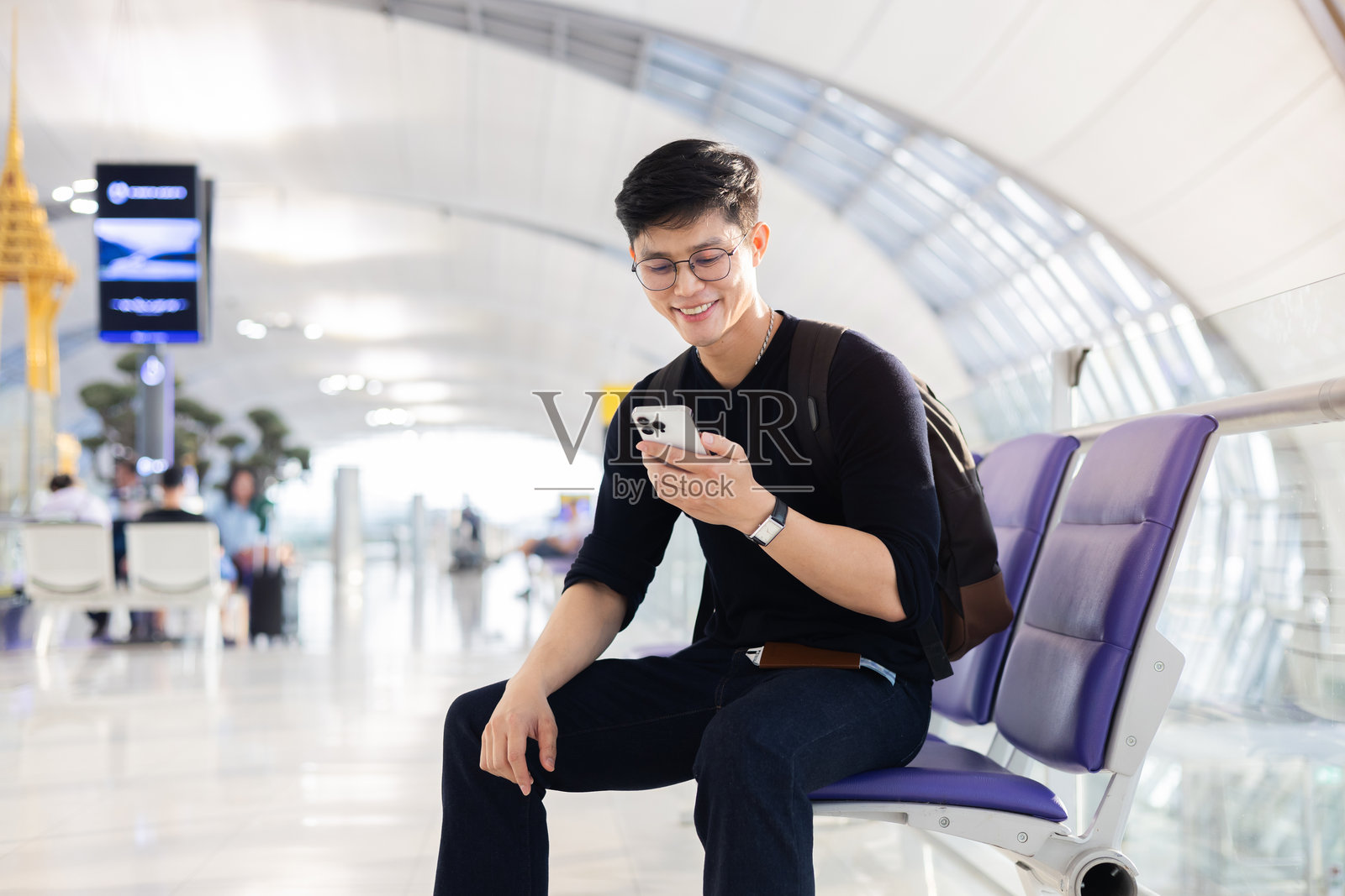 年轻的亚洲男子微笑着，手持护照和机票，坐在机场候机区使用智能手机，展现旅行和人们的生活方式概念。照片摄影图片