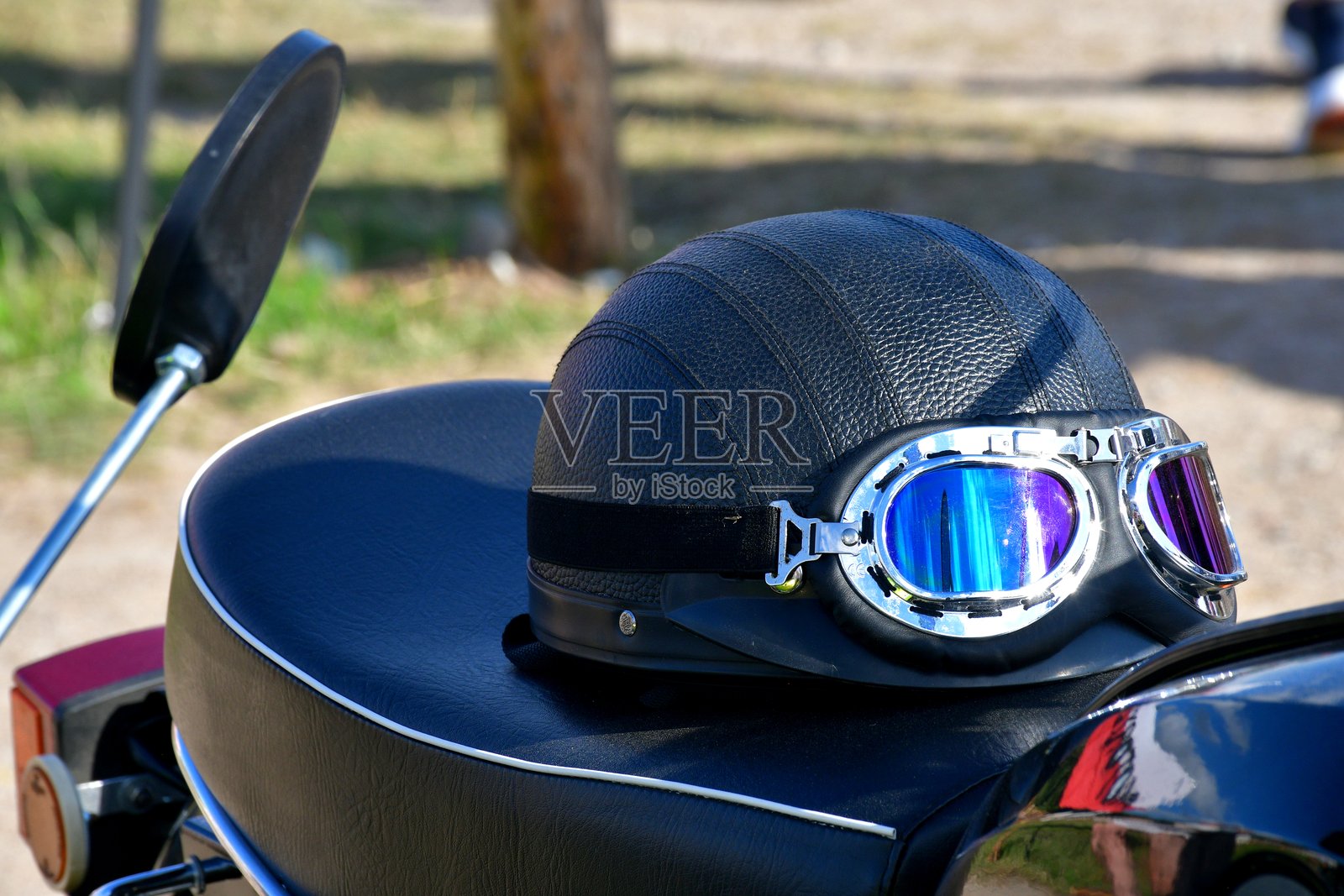 一顶坚固的皮革头盔特写，配有一副护目镜，放在复古摩托车的皮革座椅上，背景是波兰阳光明媚的夏日，旁边是一片郁郁葱葱的田野或草地。照片摄影图片