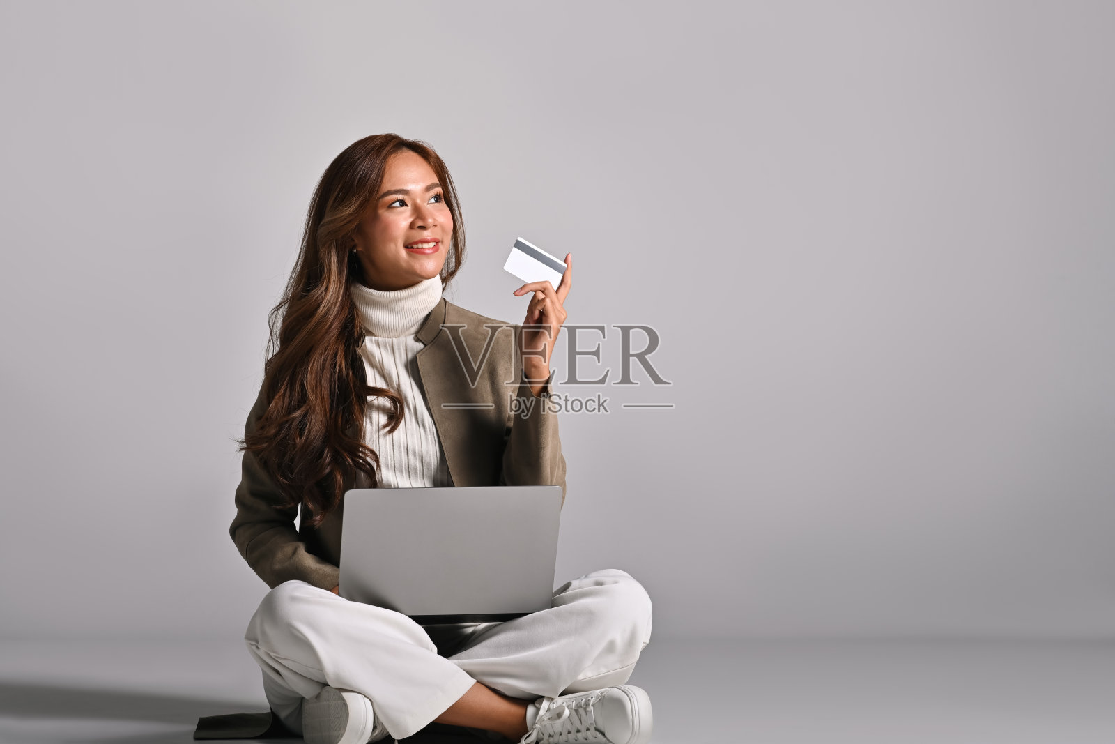 年轻女性坐在工作室里，双腿交叉，膝上放着笔记本电脑，手中拿着信用卡，面带微笑，象征着在线购物的便利和赋权，背景简洁。照片摄影图片