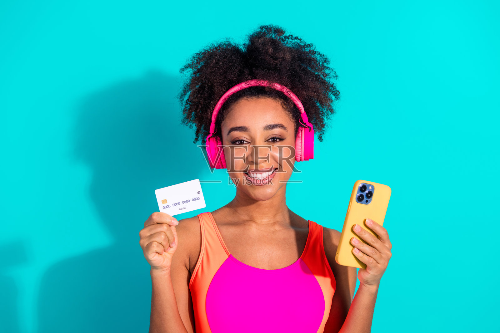 年轻快乐的女性穿着色彩斑斓的服装，手持智能手机和信用卡，展示她充满活力的风格，背景为青绿色。照片摄影图片