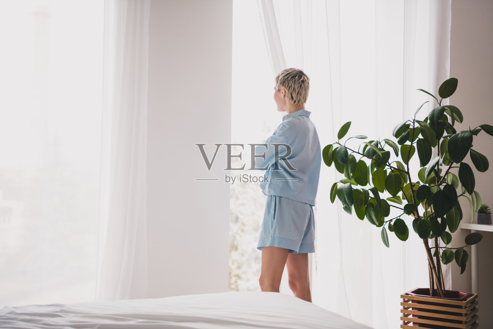 穿着蓝色睡衣的年轻女性站在明亮的卧室里，旁边有一株大型室内植物照片摄影图片