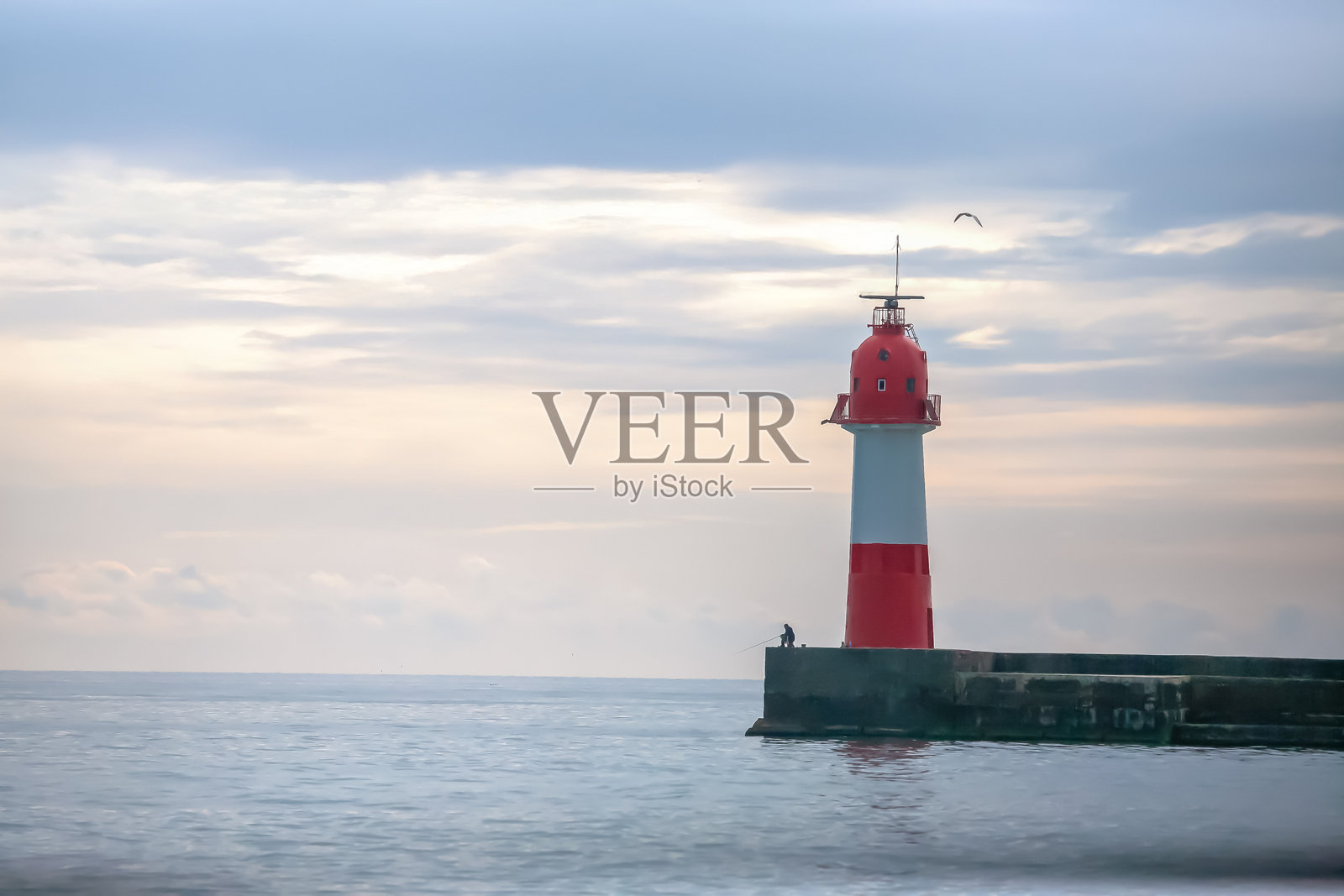 宁静的海岸场景，红白相间的灯塔矗立在码头上，一位渔夫正在抛竿，平静的海面在柔和的晨曦下显得格外迷人。照片摄影图片