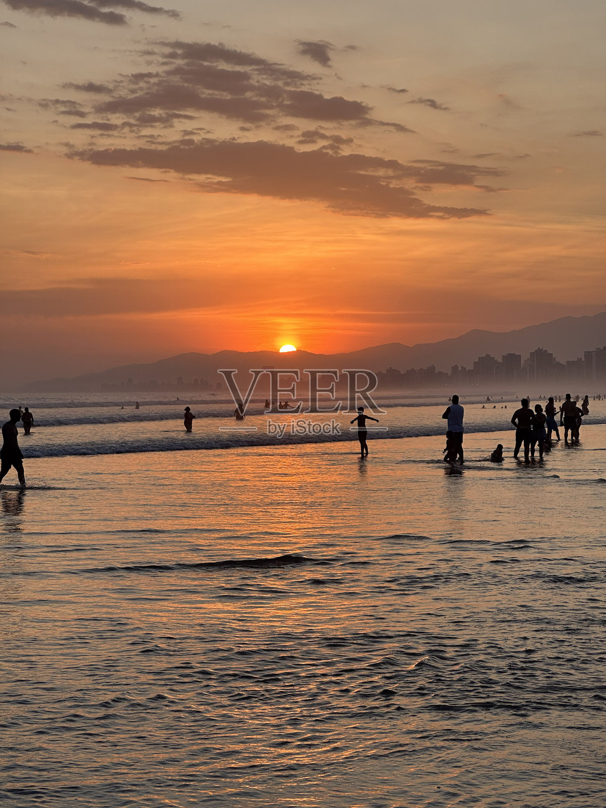 海洋上方绚丽的橙色日落,剪影中的人们享受着海洋,远处的建筑和山脉勾勒出天际线。照片摄影图片