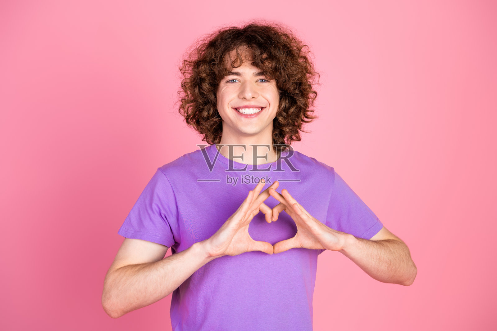 年轻男子穿着紫色T恤，卷曲的棕色头发，在鲜艳的粉色背景前做出心形手势，愉快地微笑。照片摄影图片