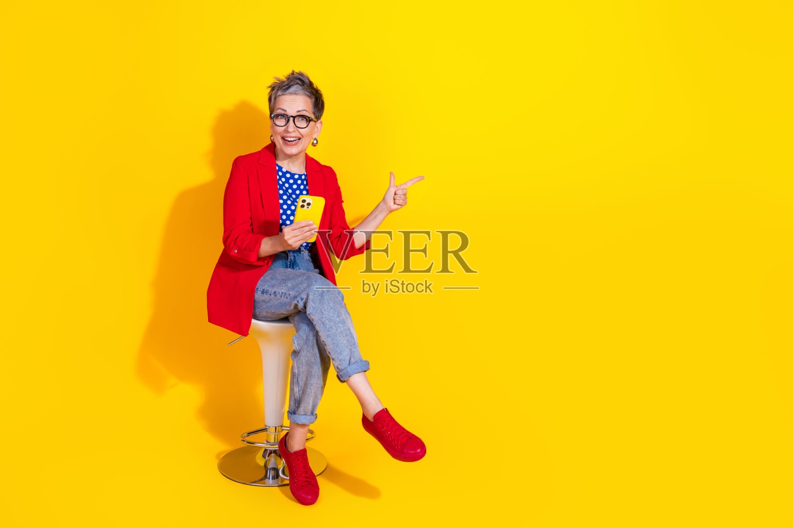 时尚的成熟的女性穿着色彩斑斓的服装，手持手机在充满活力的黄色背景前摆姿势，展现出愉快而时尚的风格。照片摄影图片