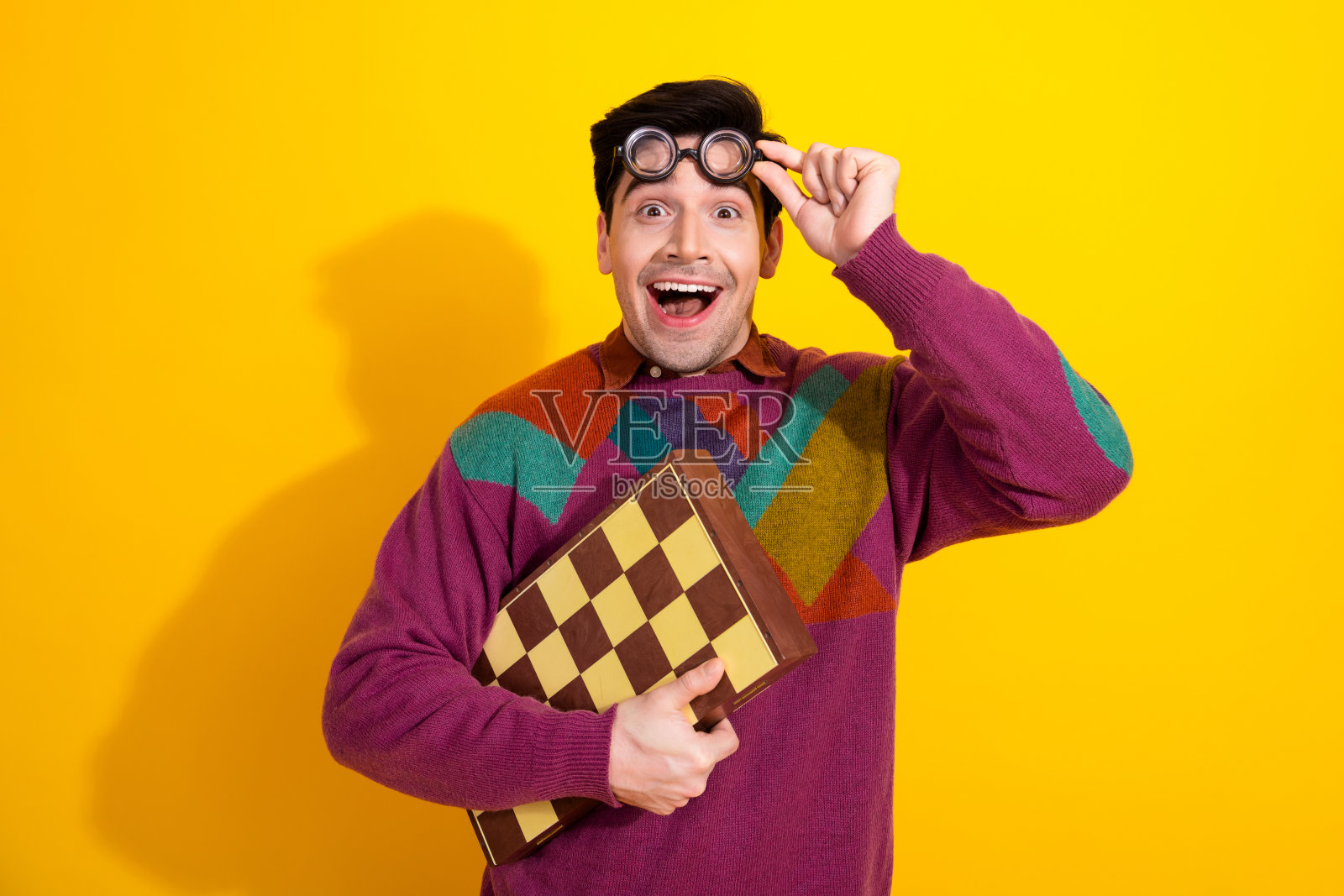 穿着五彩缤纷毛衣的快乐男人在鲜艳的黄色背景前手持棋盘，展现出惊讶和喜悦。照片摄影图片
