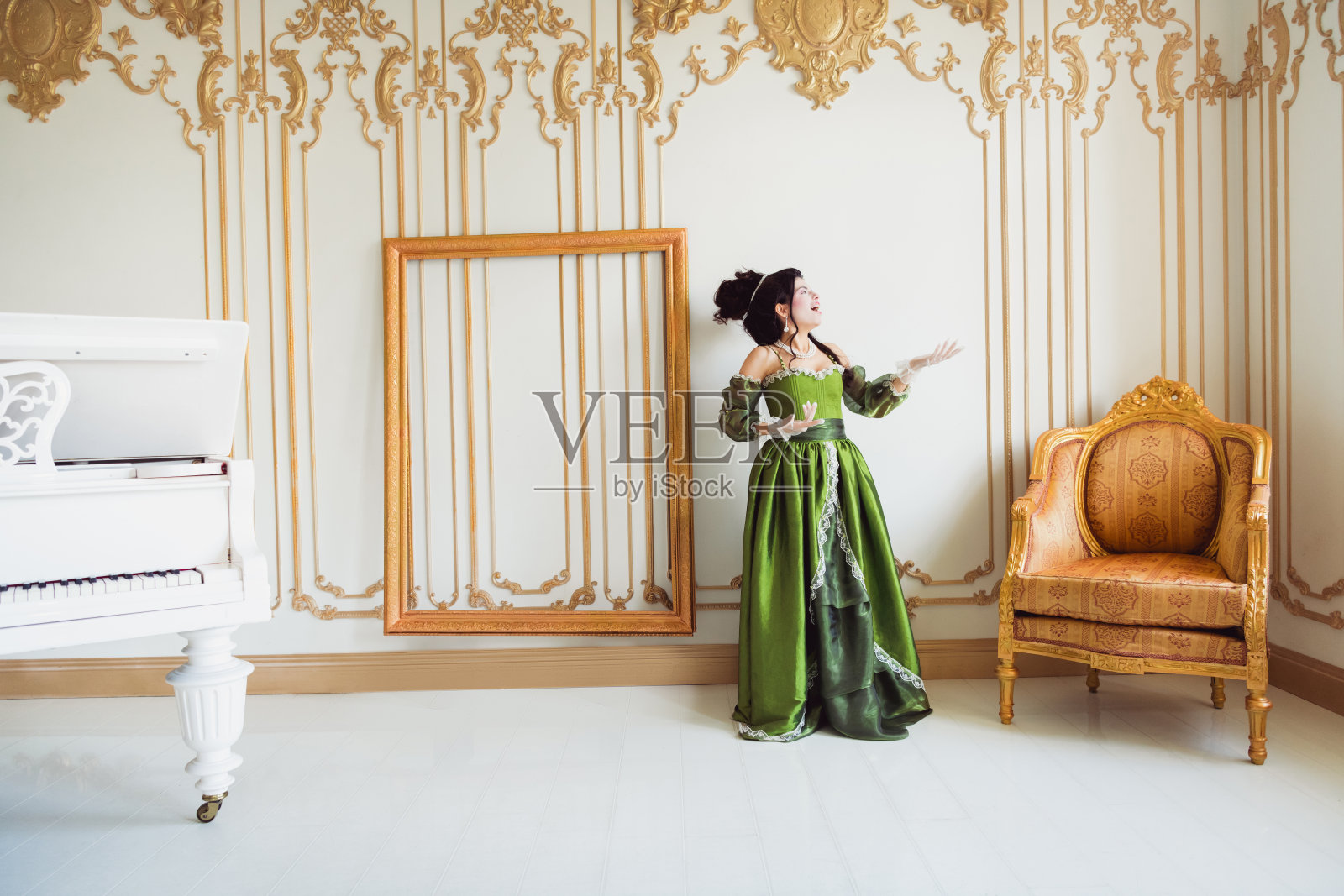 身穿绿色中世纪风格礼服的优雅女性，坐在奢华的巴洛克风格室内，周围有一把金色椅子、空框和白色大钢琴。照片摄影图片