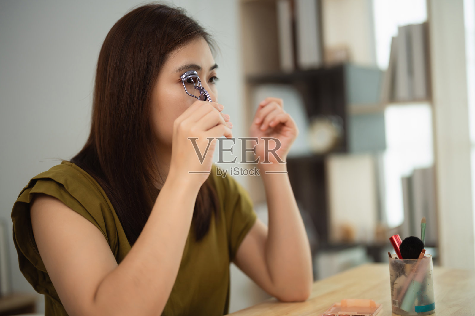 亚洲年轻女性在现代居住空间中，利用自然光和桌上的化妆工具，进行日常美容程序，使用睫毛夹。照片摄影图片
