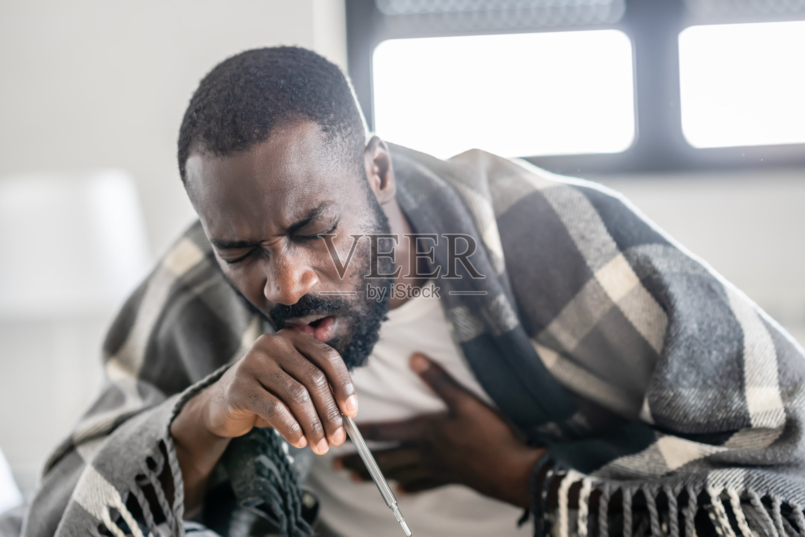 生病的男人裹着格子毛毯，坐在室内，用拳头咳嗽，看起来不舒服且疲惫。照片摄影图片