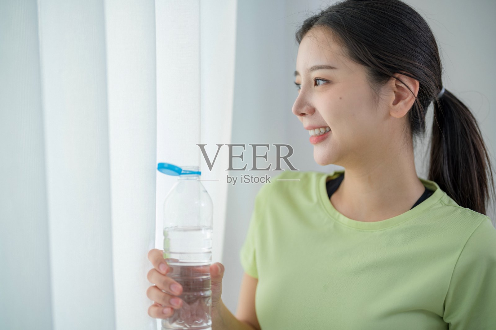 穿着绿色衬衫的女人在明亮的早晨微笑着，手里拿着水瓶，靠近窗户。照片摄影图片