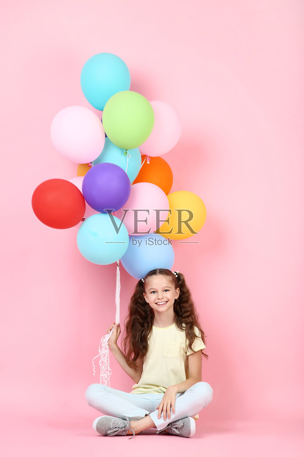 可爱的小女孩坐在粉色背景上，手里拿着彩色气球。照片摄影图片