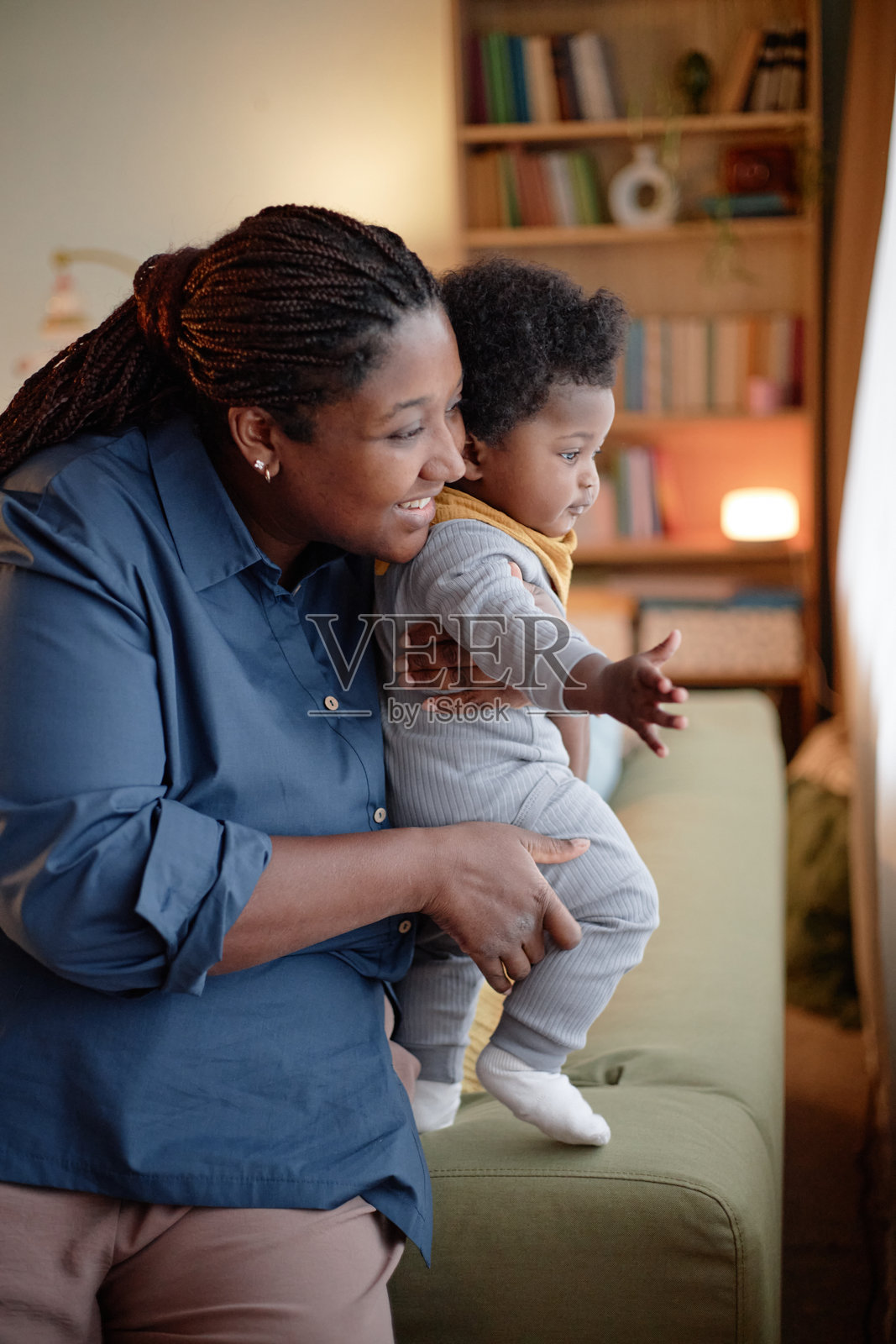 微笑的黑人母亲抱着小女孩，透过卧室的窗户看外面。照片摄影图片