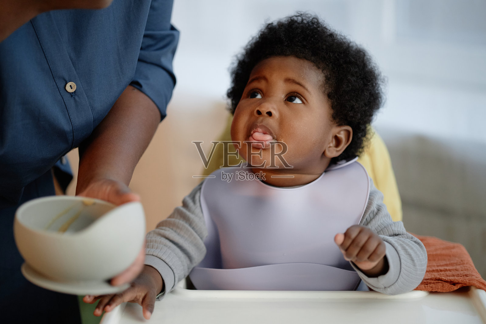 饥饿的黑肤小女孩穿着围嘴坐在高脚椅上，妈妈在室内喂她。照片摄影图片