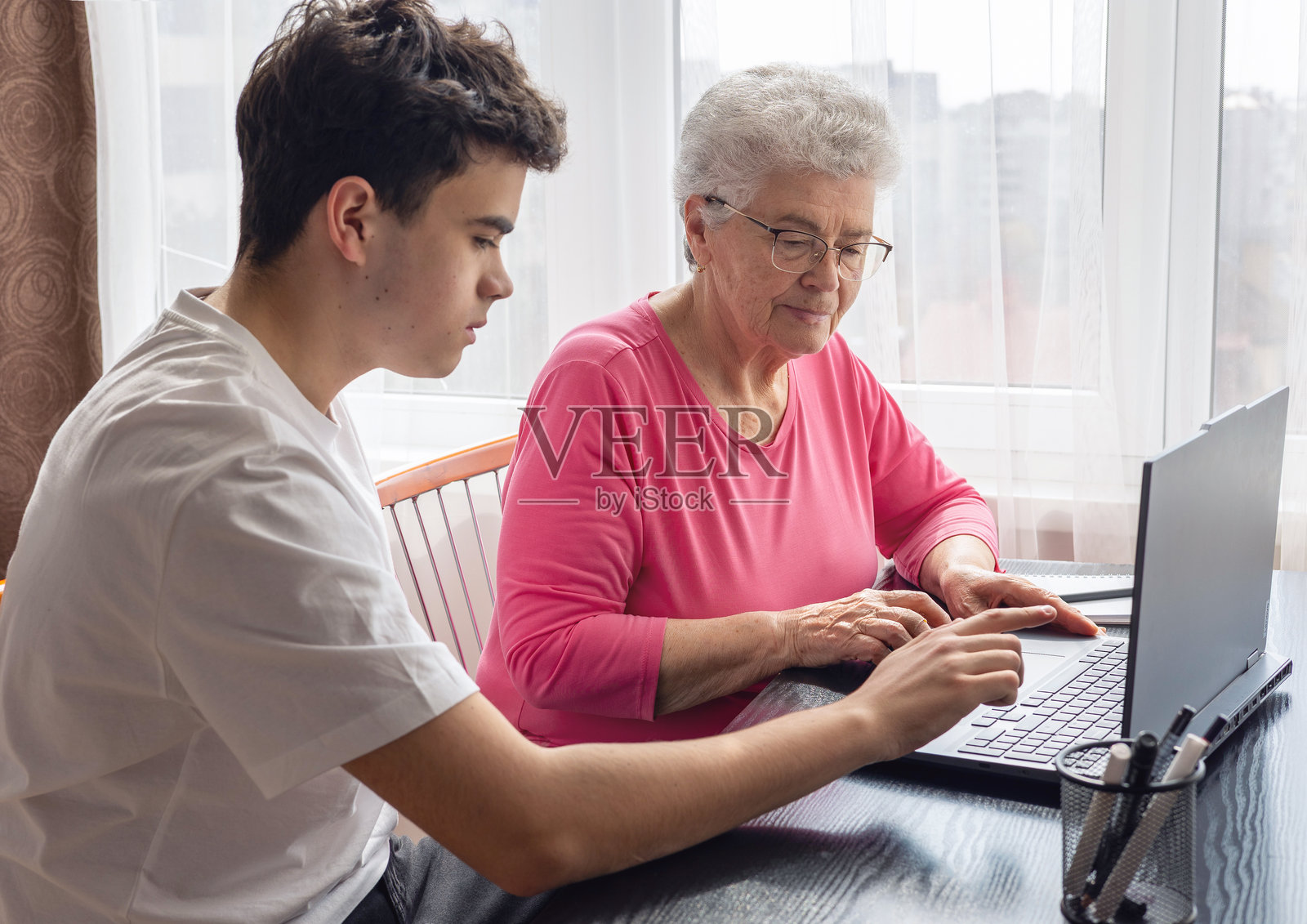孙子帮助祖母使用电脑，教授数字技能，老年女性学习科技，家庭支持，代际沟通，老年教育，在线学习辅助。老年人的数字素养。照片摄影图片