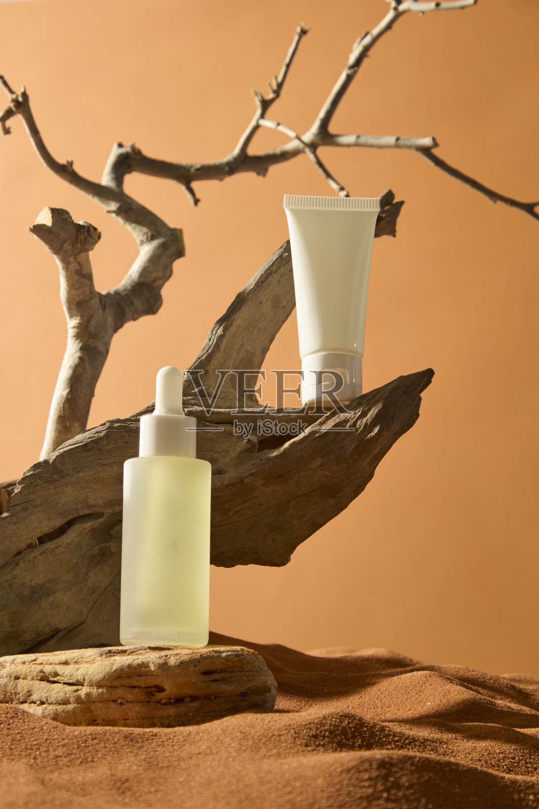 无标签的白色管子放置在干燥的木头树干上，尖端朝上。滴管瓶的精华液展示在具有凹凸边缘的天然石头上。展示背景为沙漠中的沙地。照片摄影图片
